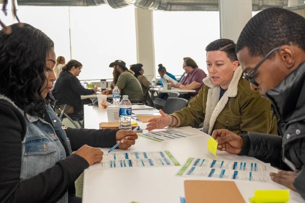 People sitting at a table having a discussion, with papers, notebooks, and water bottles in front of them, in a well-lit room with large windows.