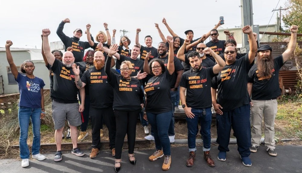 A diverse group of people posing outdoors, some with their fists raised, celebrating together, with many wearing black T-shirts that say 'Working Class of 2023 Candidate'.