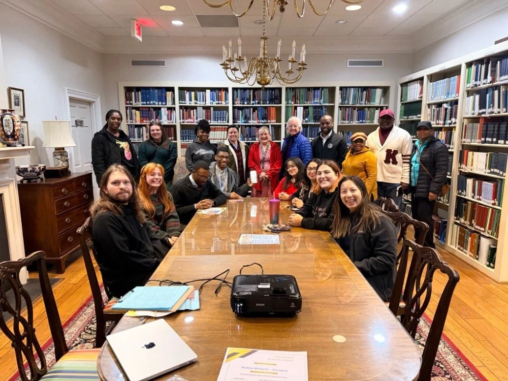 Wayne County Young Democrats in a library gathered around a long table, smiling for a photo, with bookshelves and a chandelier in the background.