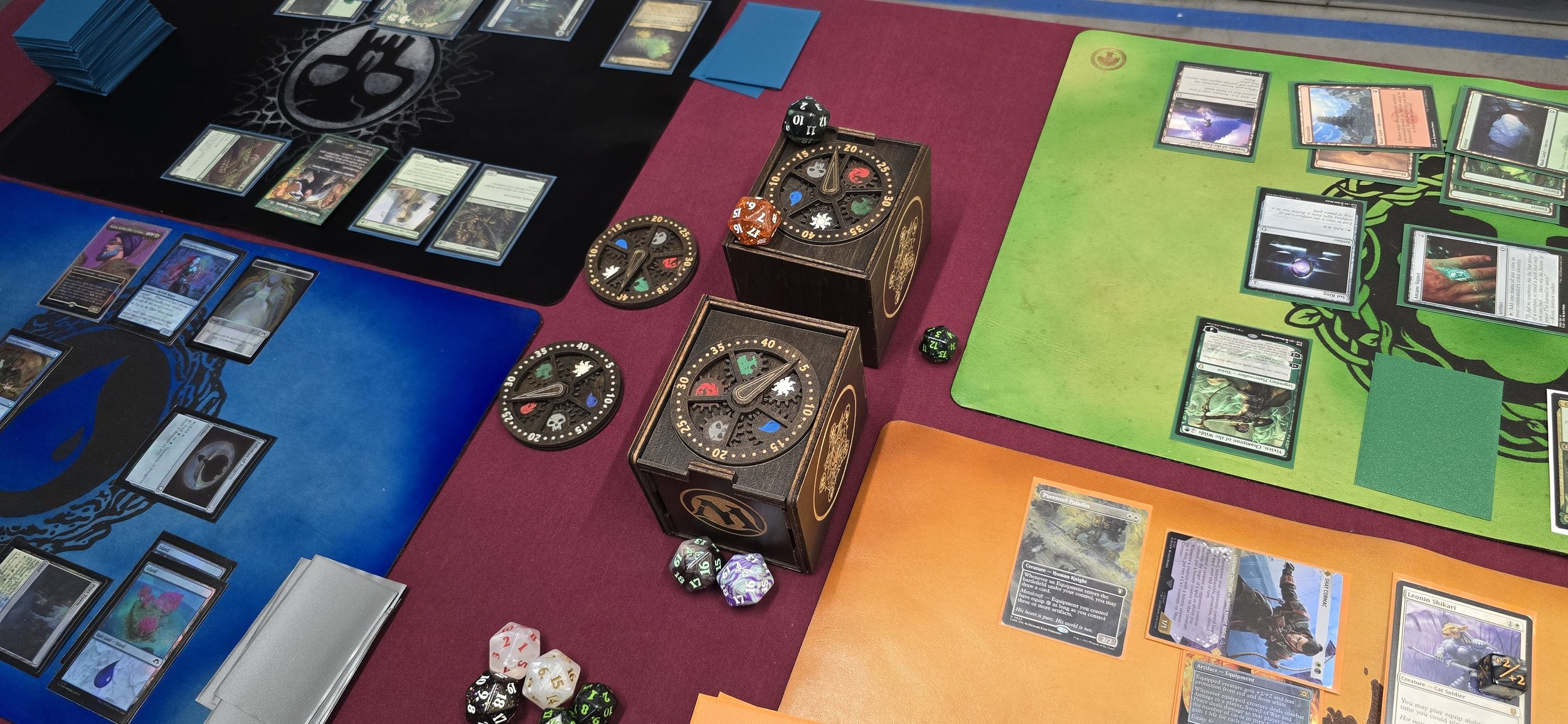 This is an overhead image of a Magic: The Gathering game in progress. It features two deck boxes, two life counters, and four leather playmats on display.
