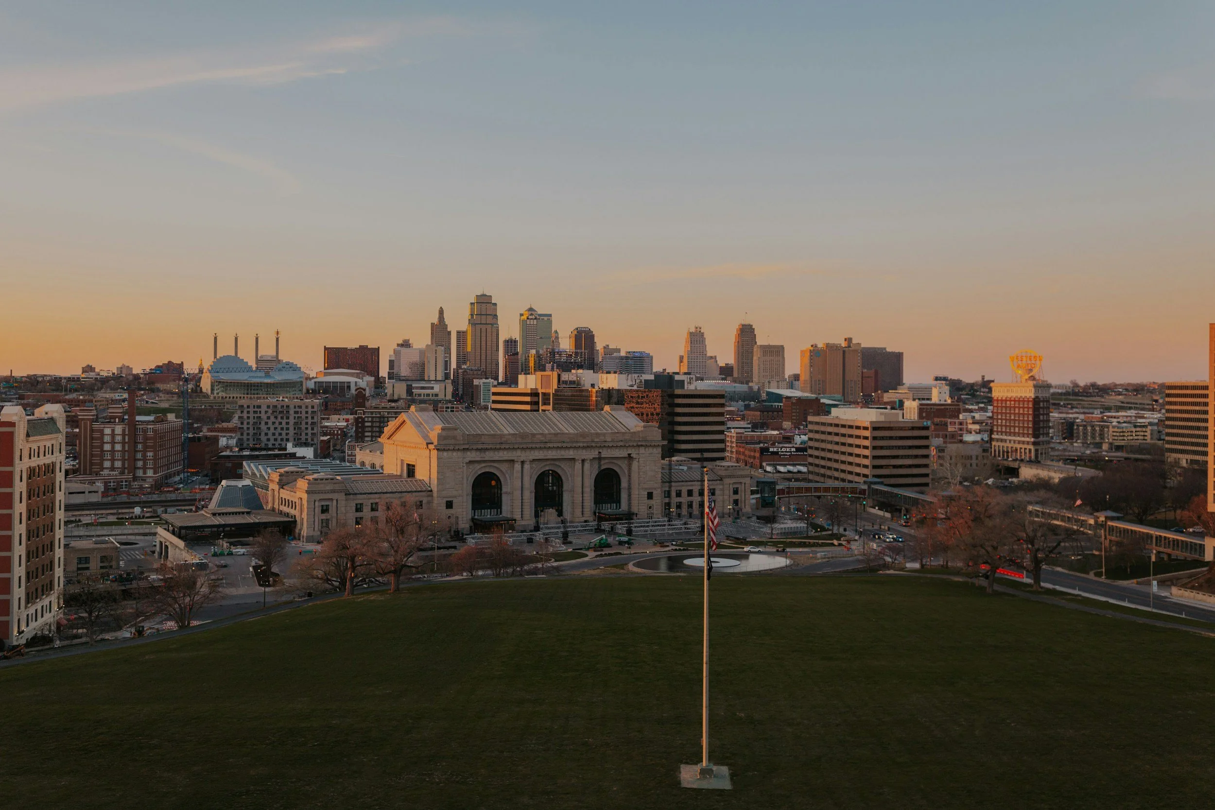 Sunset skyline of Union Station in Kansas City, including a distinctive building with a dome, and a large American flag in the foreground on a grassy area.