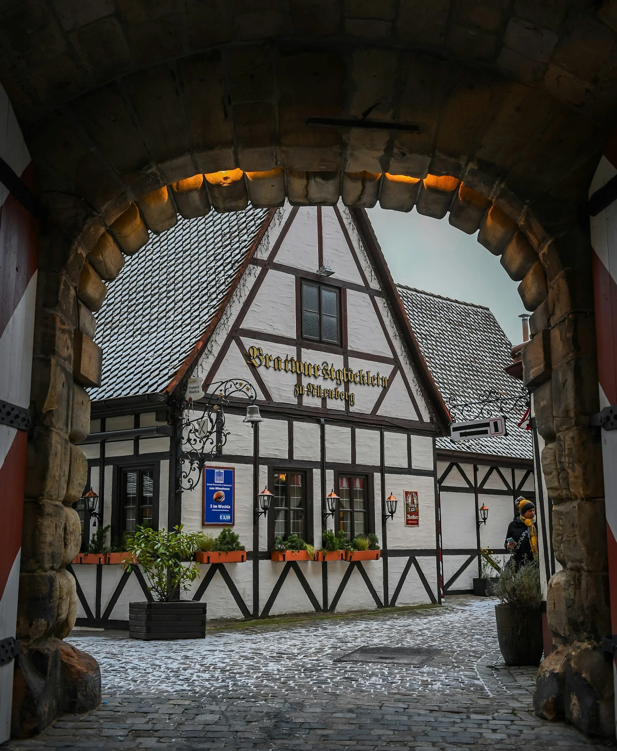 View of a quaint European-style building with white walls and dark wooden beams, decorated with hanging lanterns, seen through a stone archway in a cobblestone courtyard with a person walking and plant containers.
