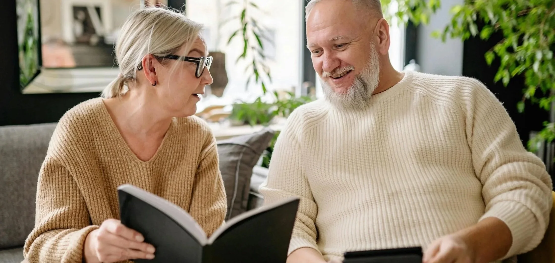 A woman and a man sitting on a couch, smiling, looking at a book and a tablet together in a cozy, well-lit living room with plants around them.