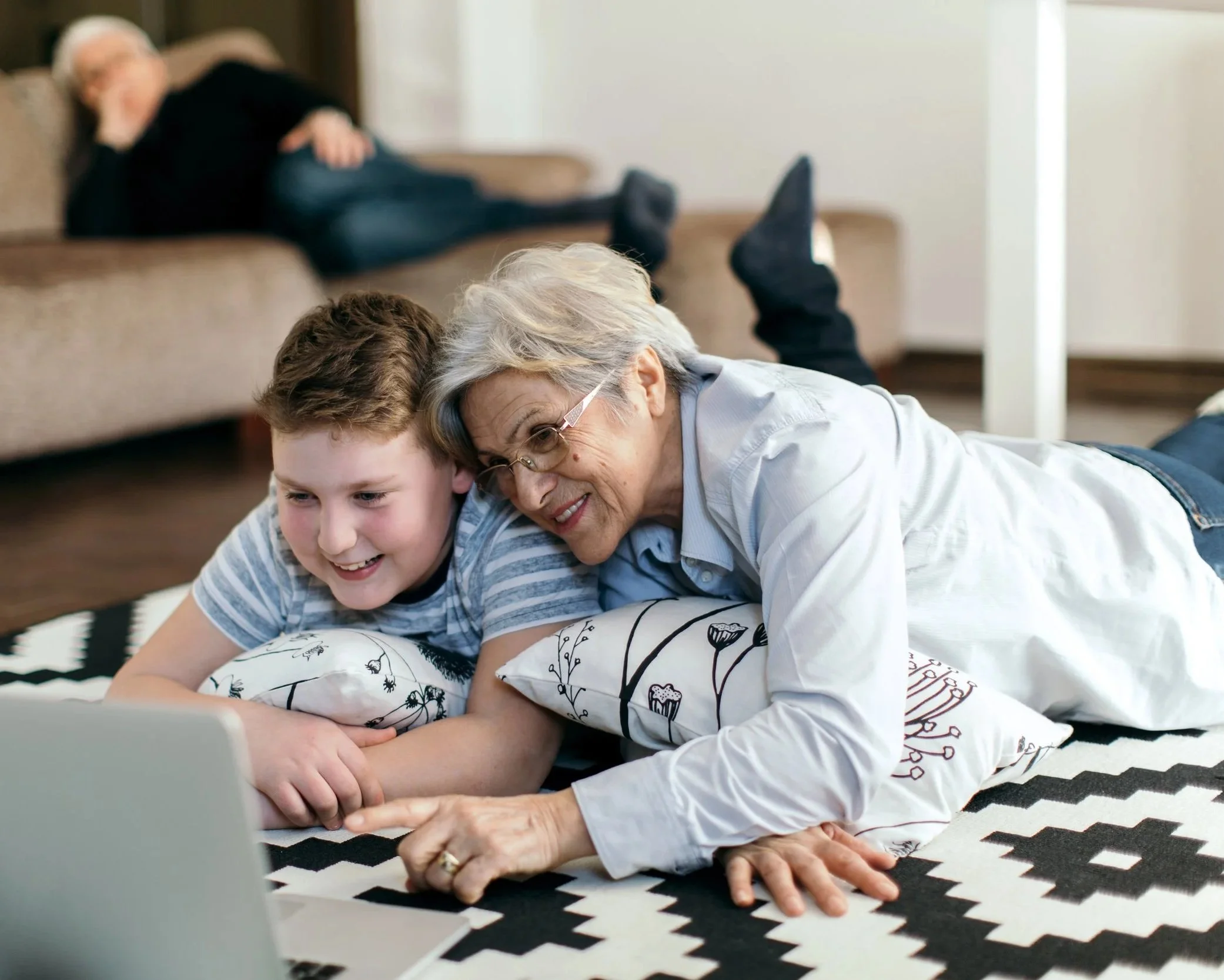 A smiling elderly woman and a young boy lying on a striped black-and-white rug, looking at a laptop, with a blurred elderly person sleeping on a sofa in the background.