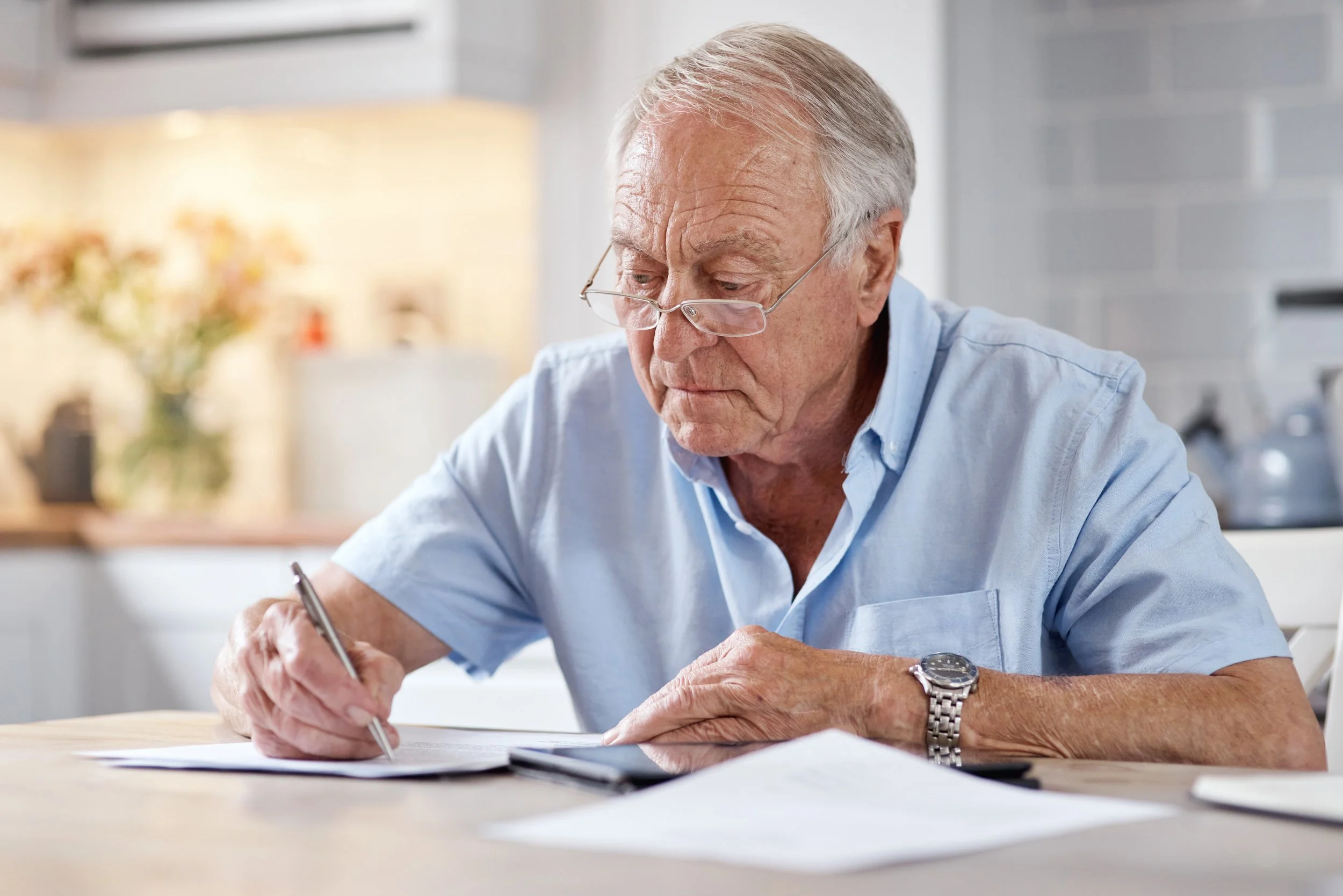An elderly man with gray hair and glasses, wearing a light blue shirt, sits at a wooden table, writing on some papers with a pen, in a bright, modern kitchen.