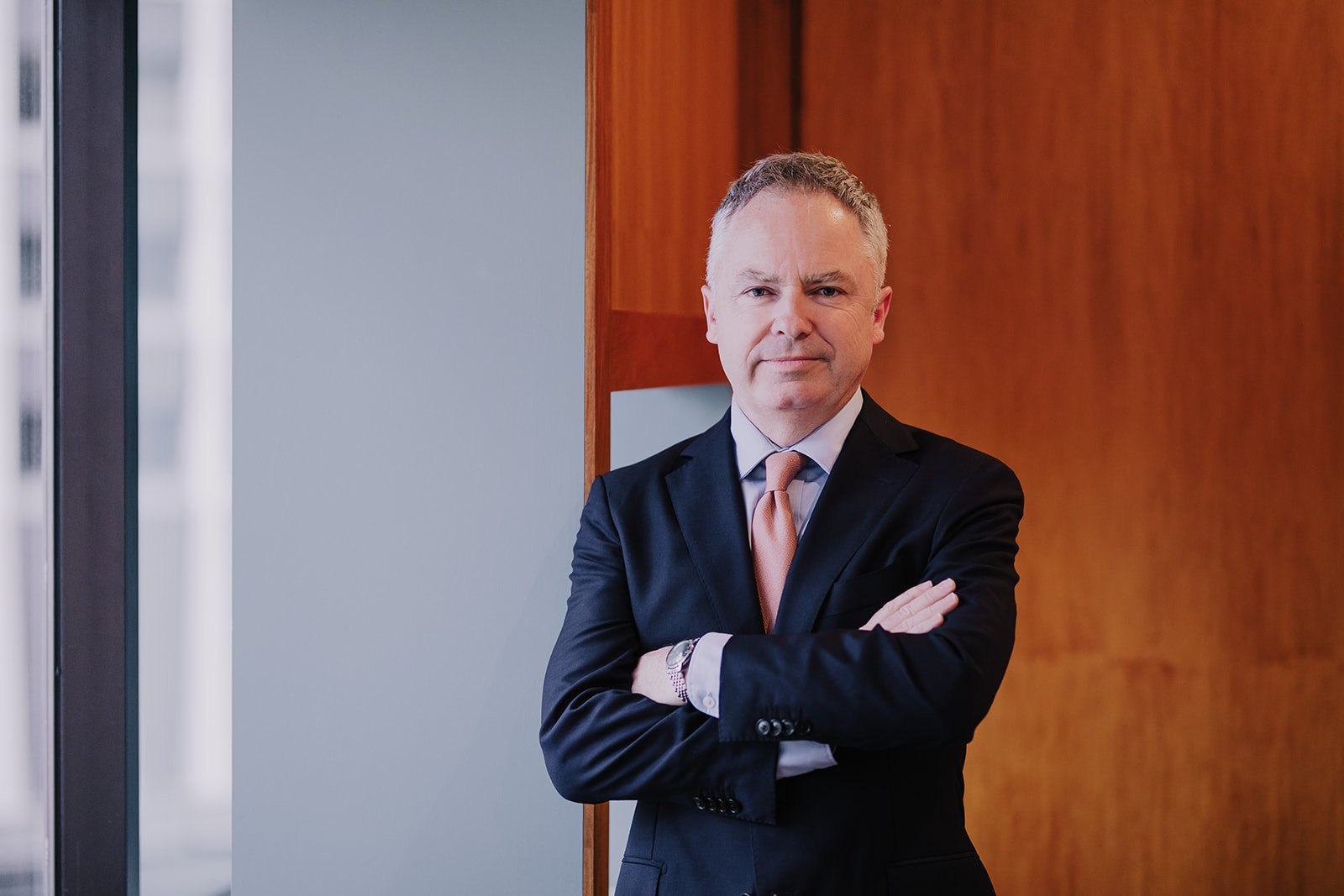 A confident middle-aged man in a suit with crossed arms, standing indoors against a wood-paneled wall with a window nearby.