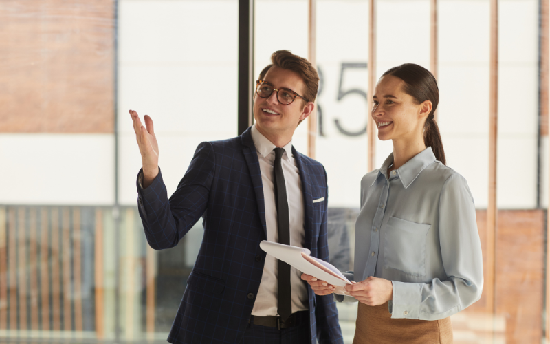 Two professionals, a man in a suit and glasses and a woman in a blouse, are talking and smiling in a modern office or conference area.