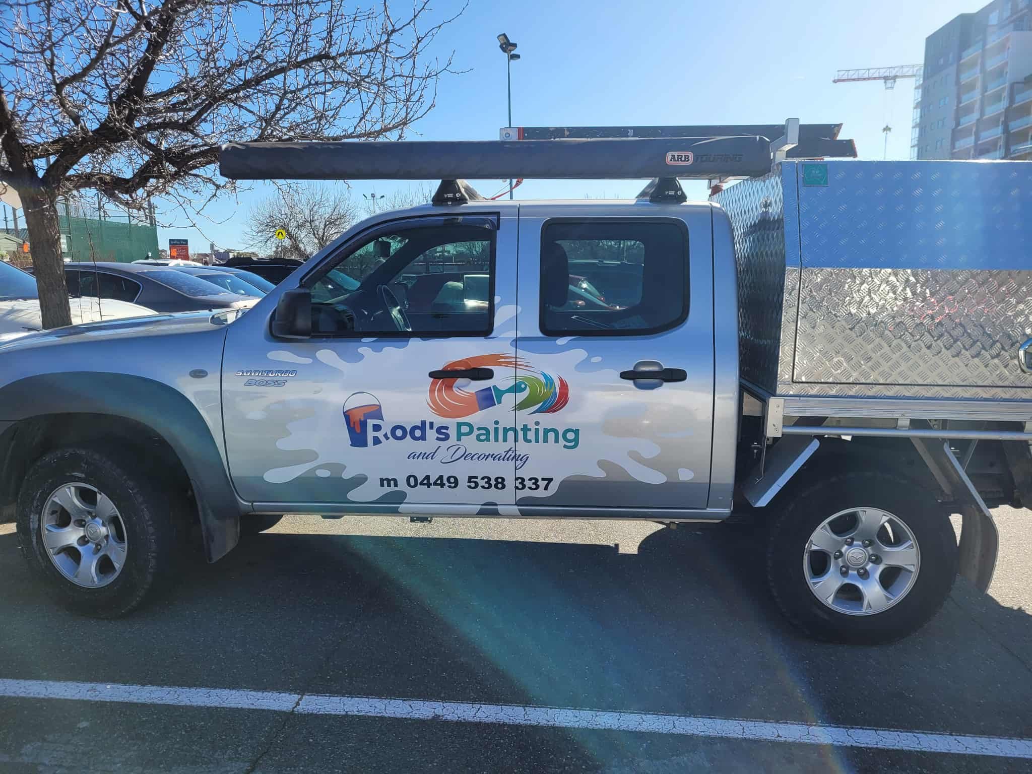 A silver work vehicle with signage for 'Rod's Painting and Decorating,' including a colorful paintbrush logo and phone number, parked in a lot with other cars under a tree.