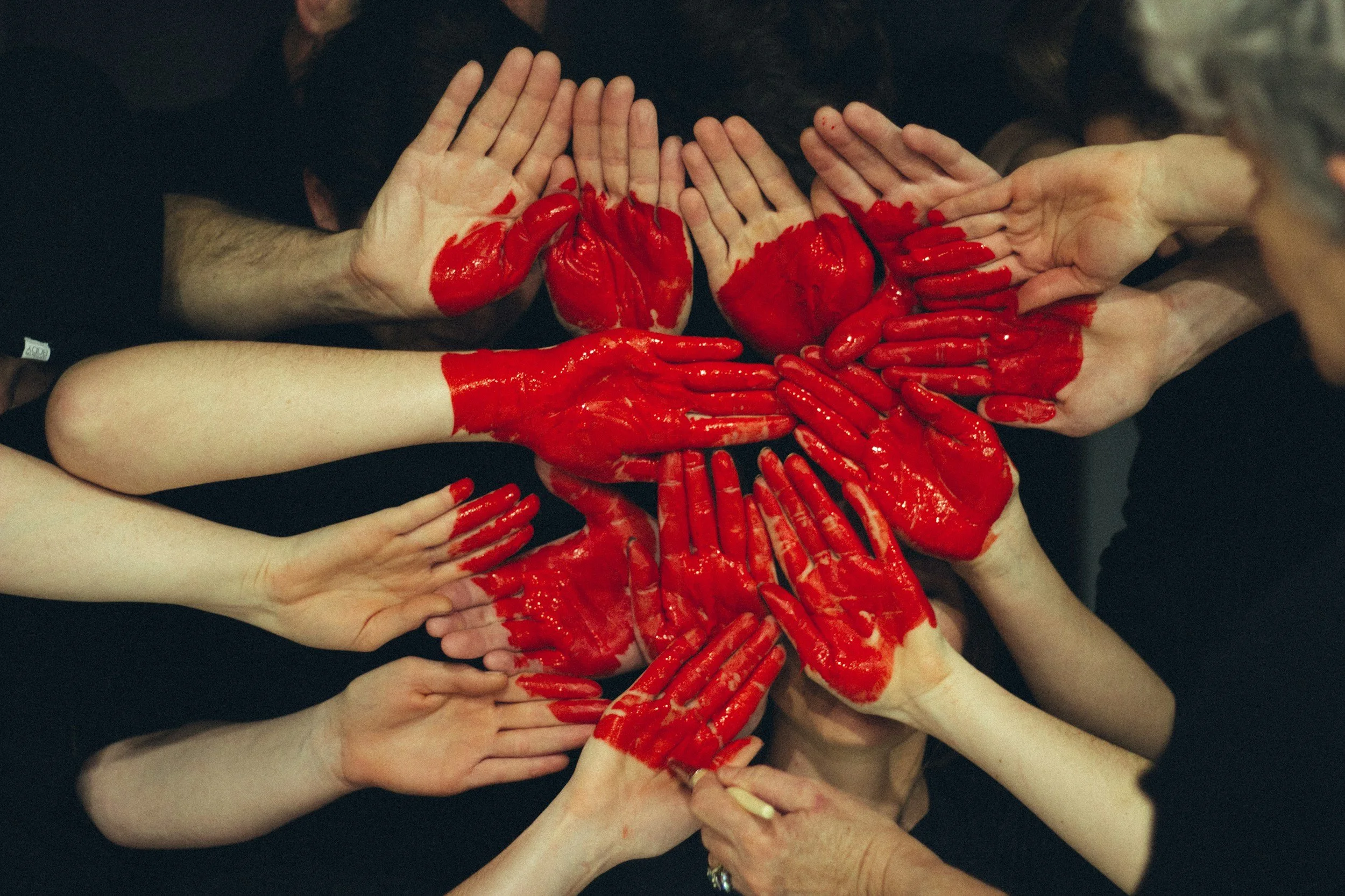 Multiple hands with red paint covering the palms and fingers, forming a layered, collective display against a dark background.
