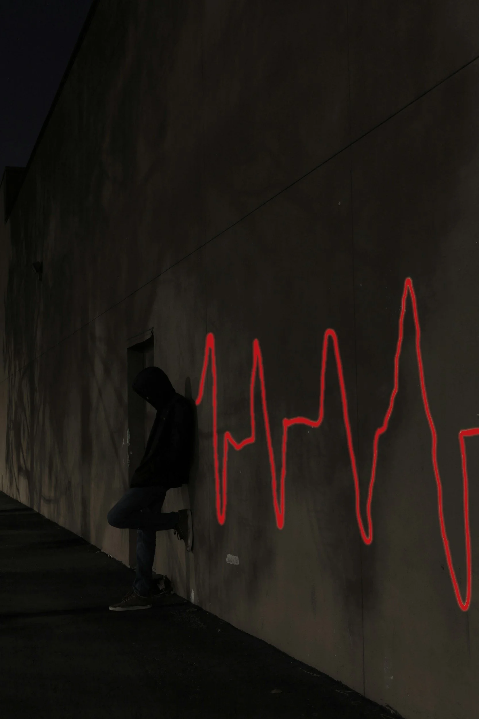 Silhouette of a person leaning against a wall at night with a red neon heartbeat line on the wall.