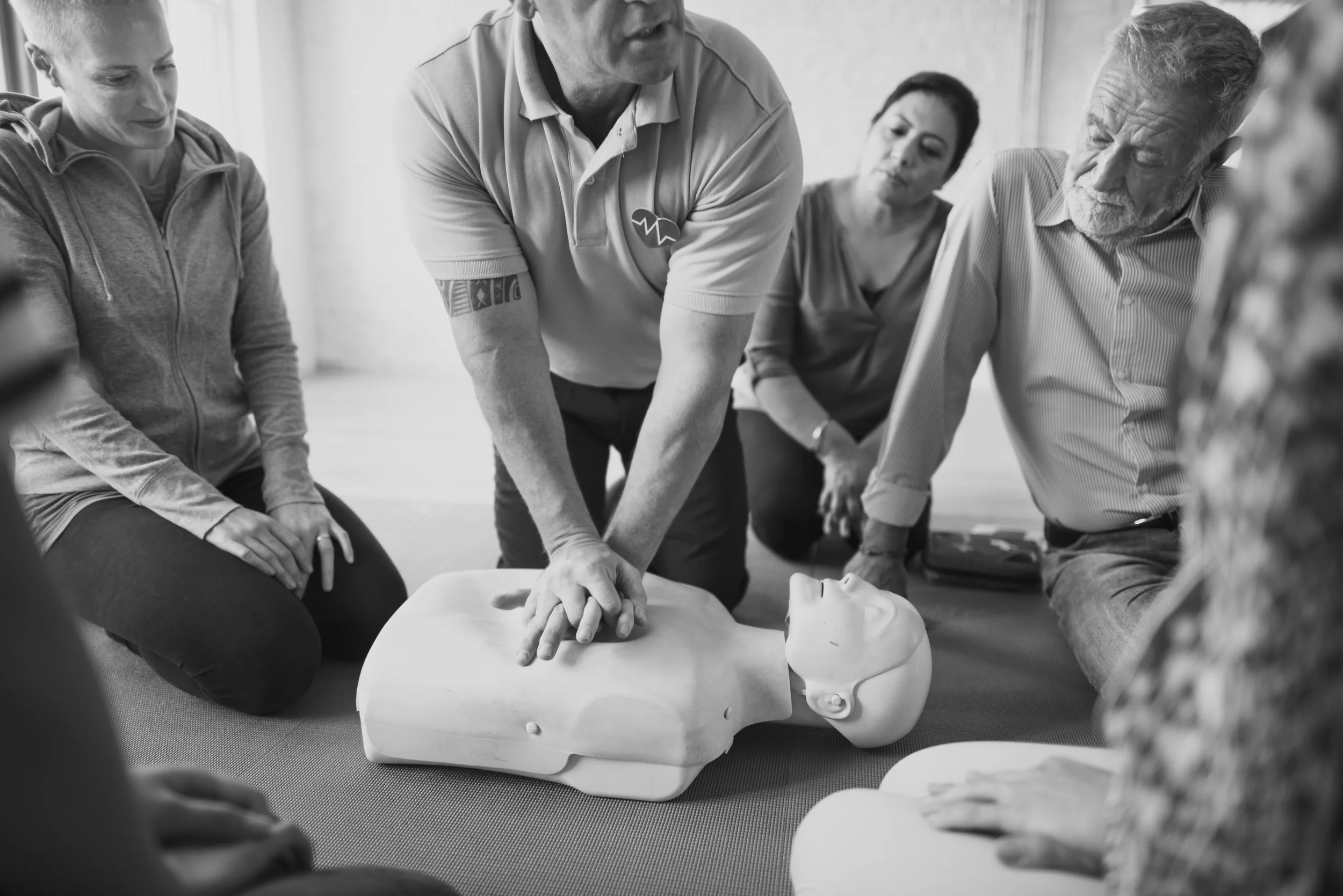 A group of people participating in a CPR training class, practicing chest compressions on a mannequin, with one person demonstrating and others observing.