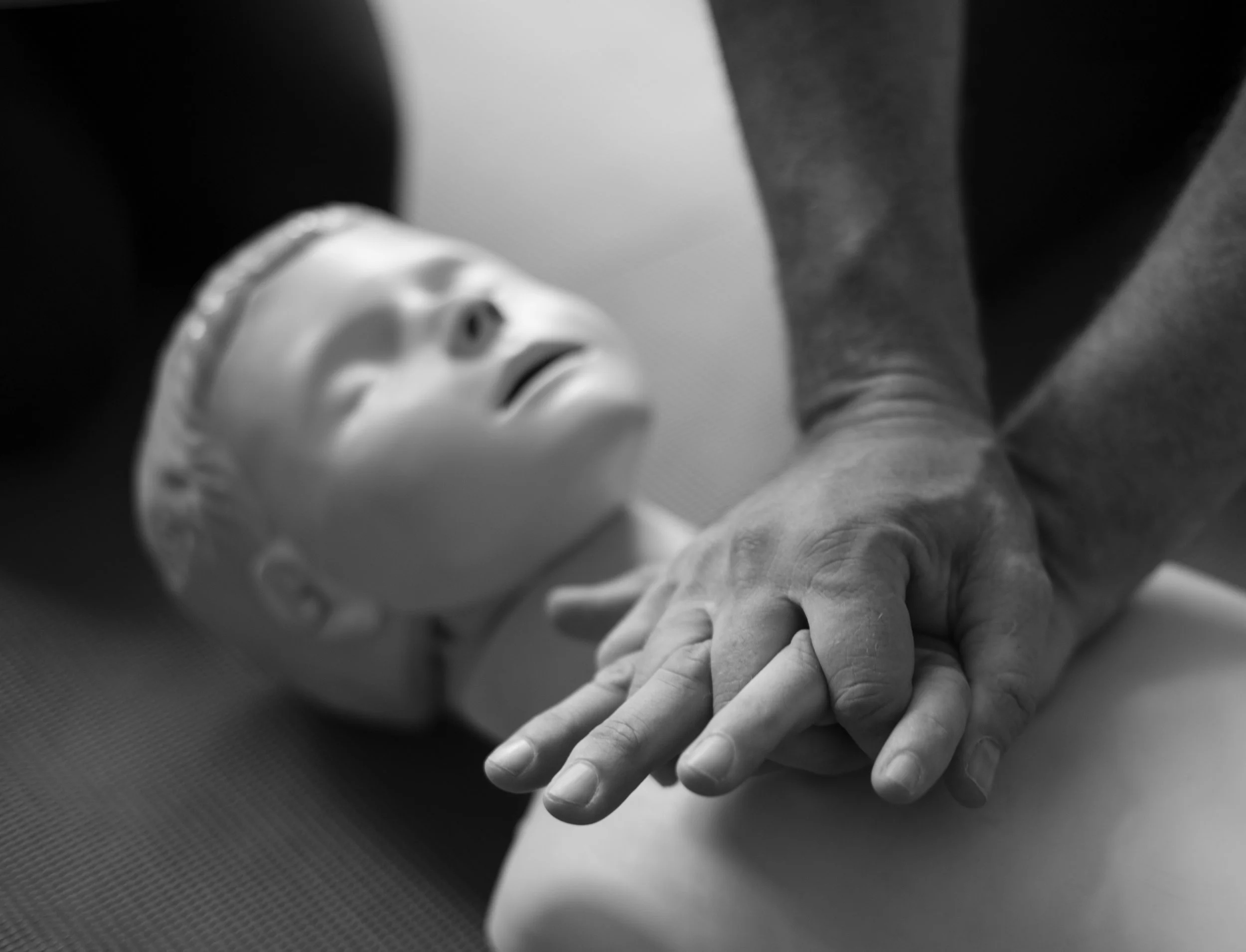 A close-up of elderly hands holding a person's hand during a CPR training session, with a mannequin with a distressed facial expression in the background.