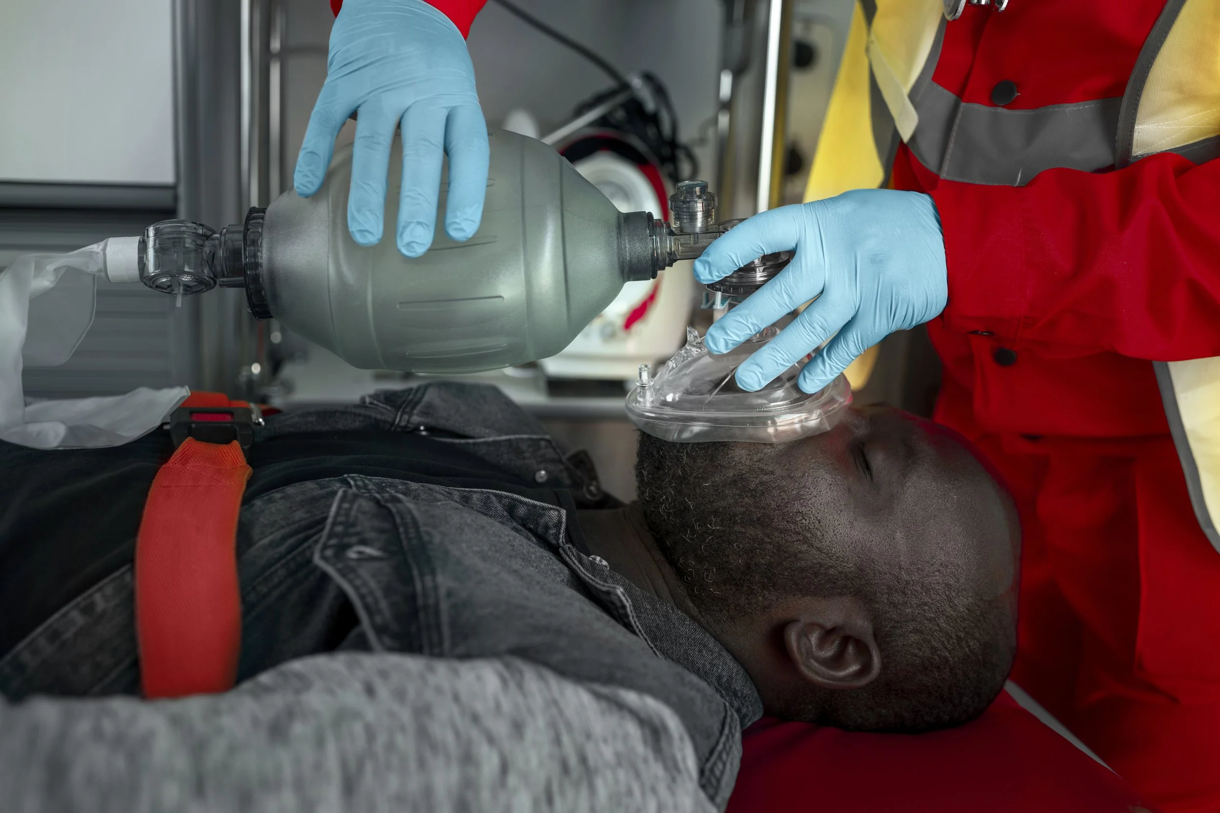 Emergency medical personnel administering oxygen to a man lying on a stretcher, using a mask connected to a bag valve mask, in a clinical setting.