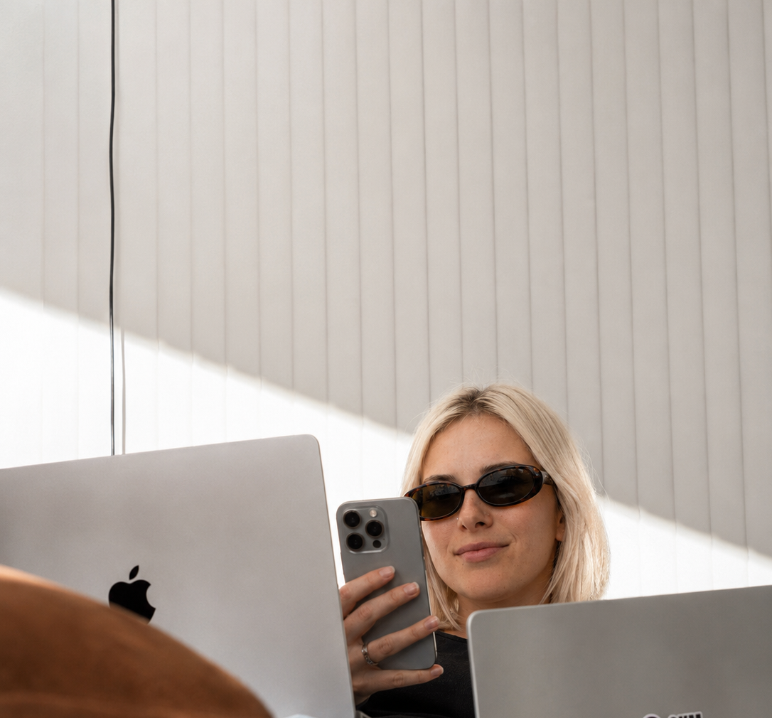Sammy with blonde hair wearing sunglasses, sitting at a desk with laptops, holding a smartphone in Downtown kelowna.