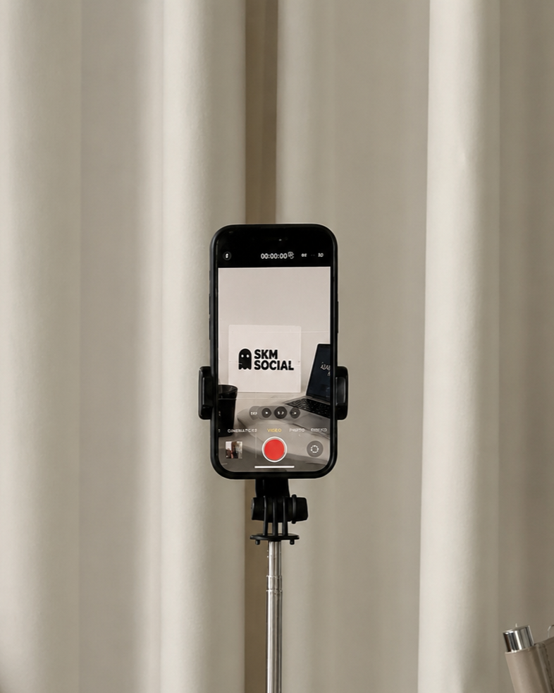 A smartphone mounted on a selfie stick recording a video of a sign that says 'SKM SOCIAL' with a ghost logo and a desk setup in the background.