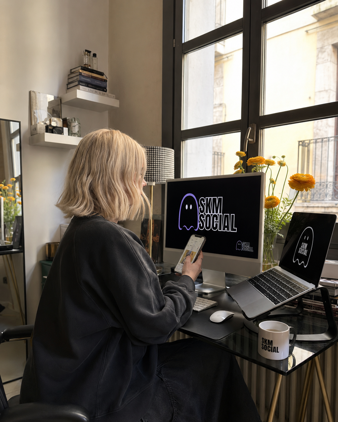 A woman with blonde hair working at a cluttered desk in a home office, looking at her phone, with a computer monitor, laptop, and SKM Social branded mug on the desk. There are yellow flowers on the windowsill and shelves with books and decorative items behind her.