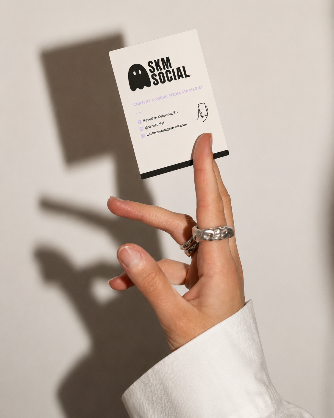 A person holding a business card with a shadow cast on a light-colored wall. The card displays contact information and social media handles for 'SKM SOCIAL,' a content and social media strategist based in Kelowna, BC.