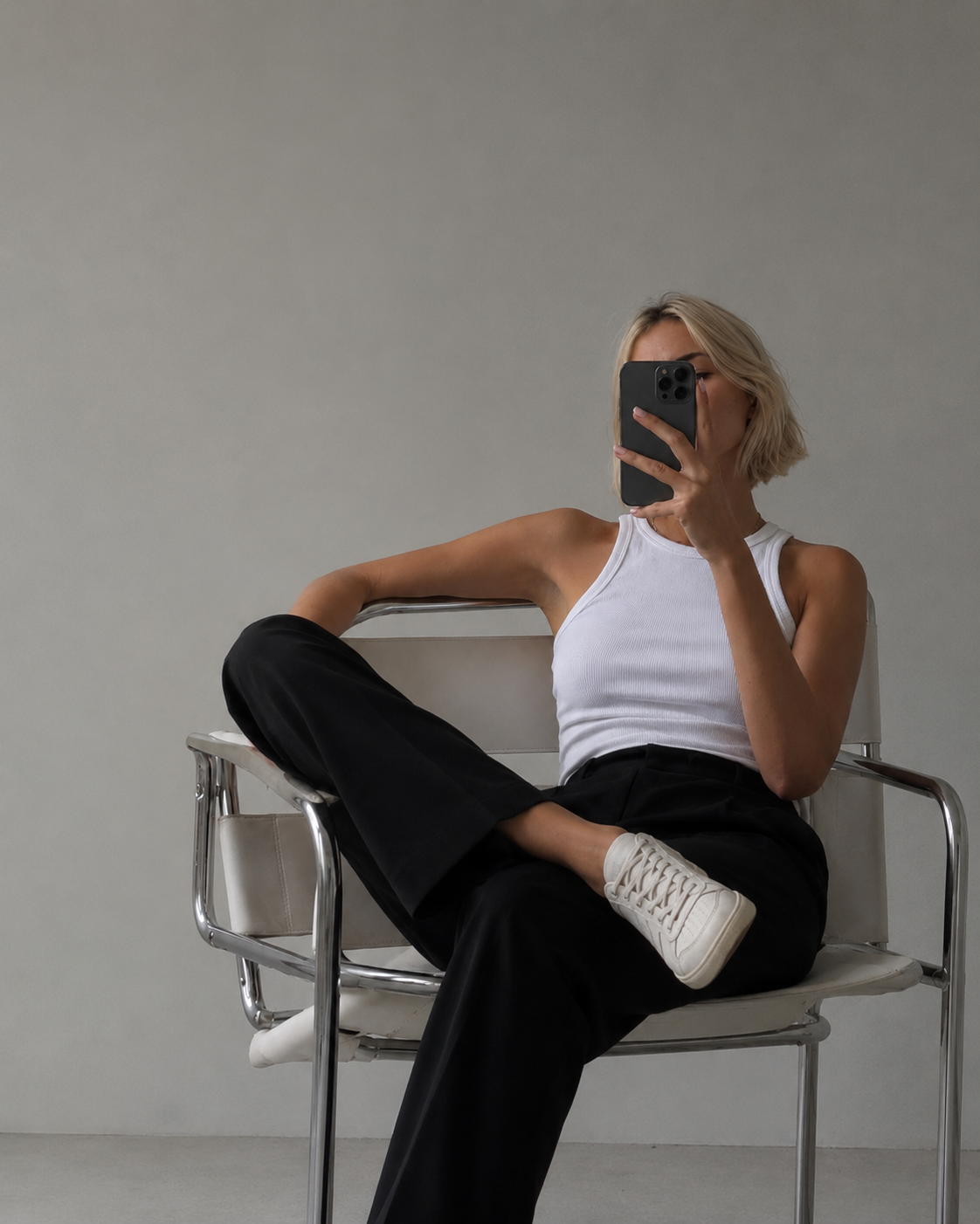 SKM SOCIAL sitting on a modern white chair, taking a mirror selfie with her phone. She is wearing a white tank top, black pants, and white sneakers, with one leg crossed over the other. The background is plain and light-colored.