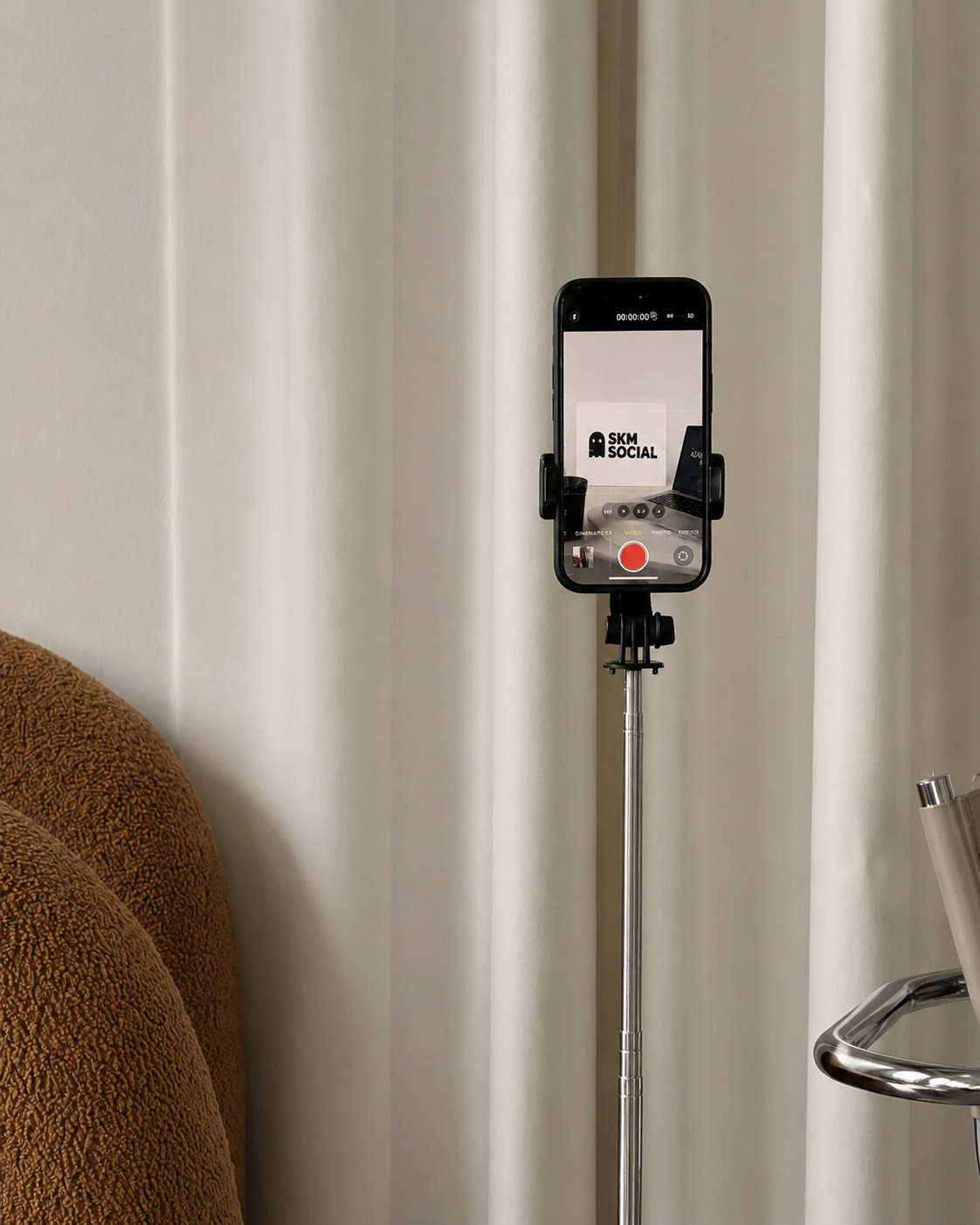 A smartphone mounted on a stand, recording a white sign with black text that reads 'SKM SOCIAL,' with a laptop and a black object behind it, against a background of beige curtains.