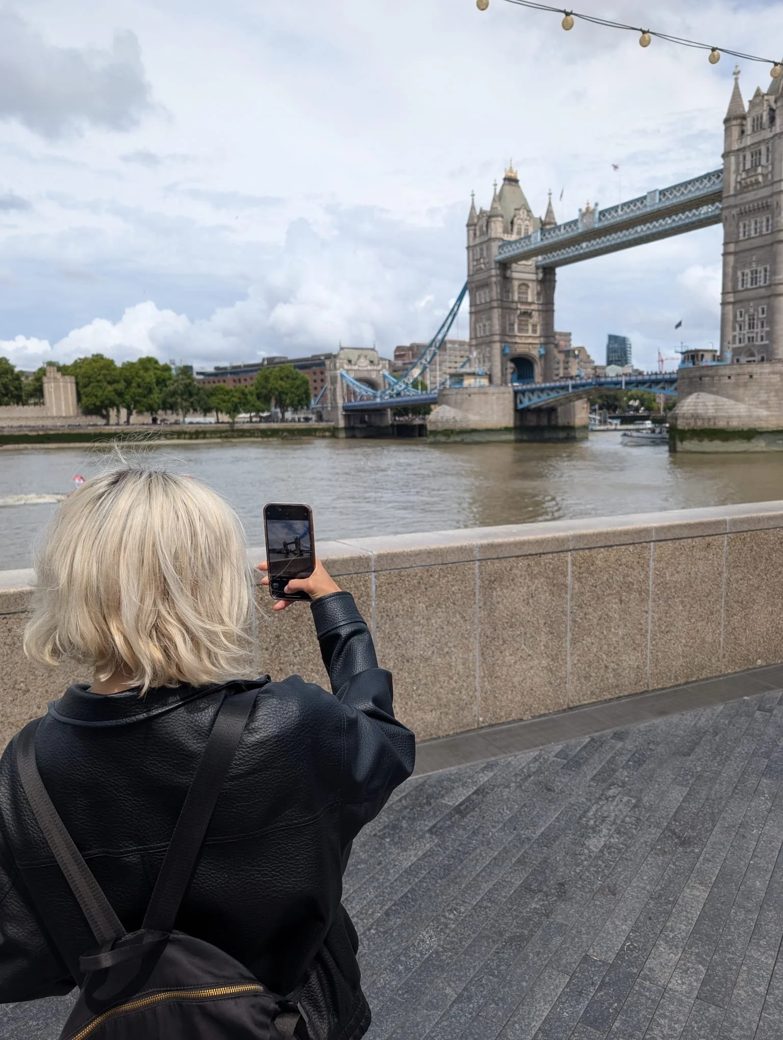 SKM SOCIAL in a black jacket is taking a photo of the Tower Bridge in London over the Thames River on a cloudy day.