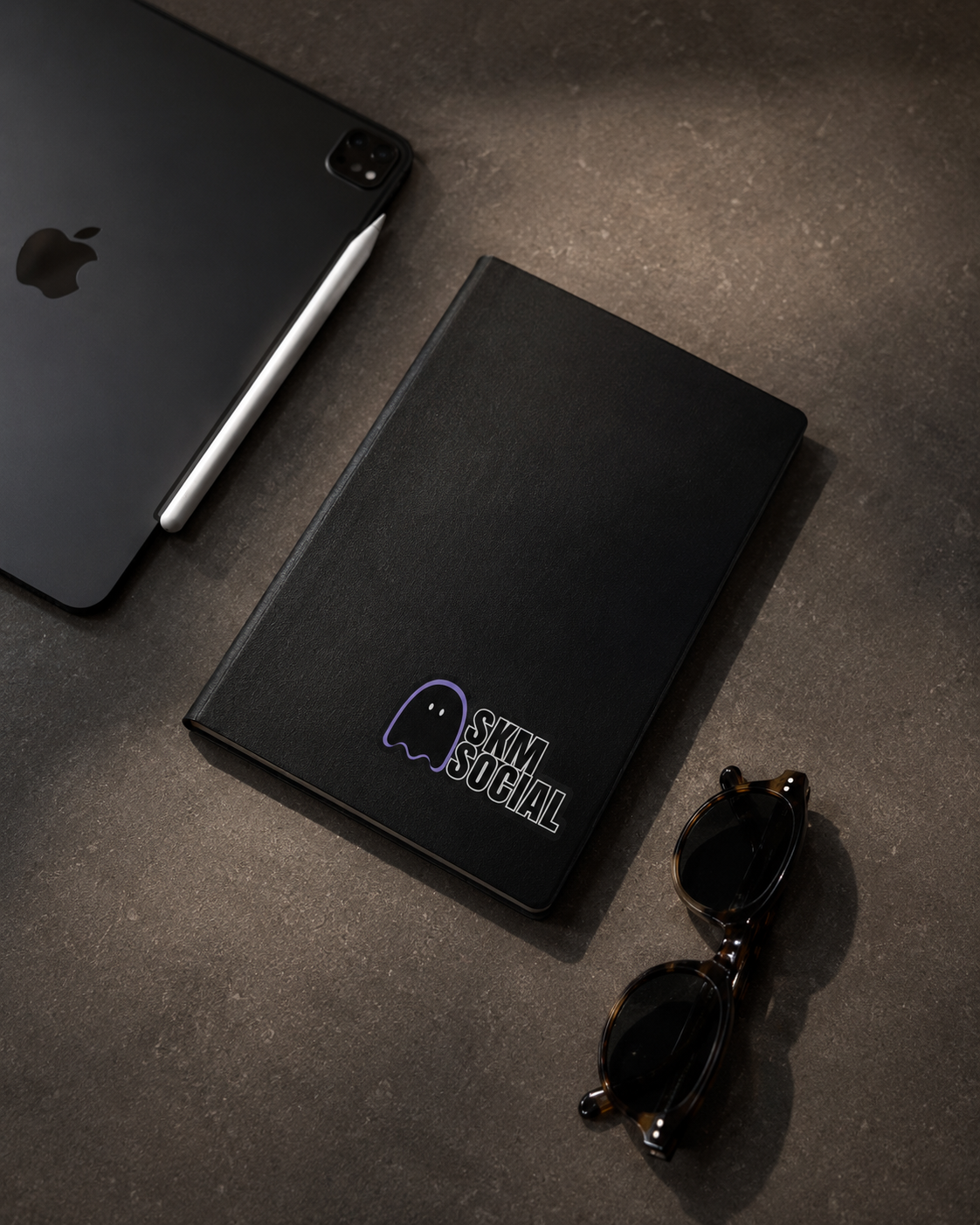A black iPad with a white stylus, a black notebook with a 'Skm Social' ghost logo, and a pair of tortoise-shell sunglasses are arranged on a dark textured surface.