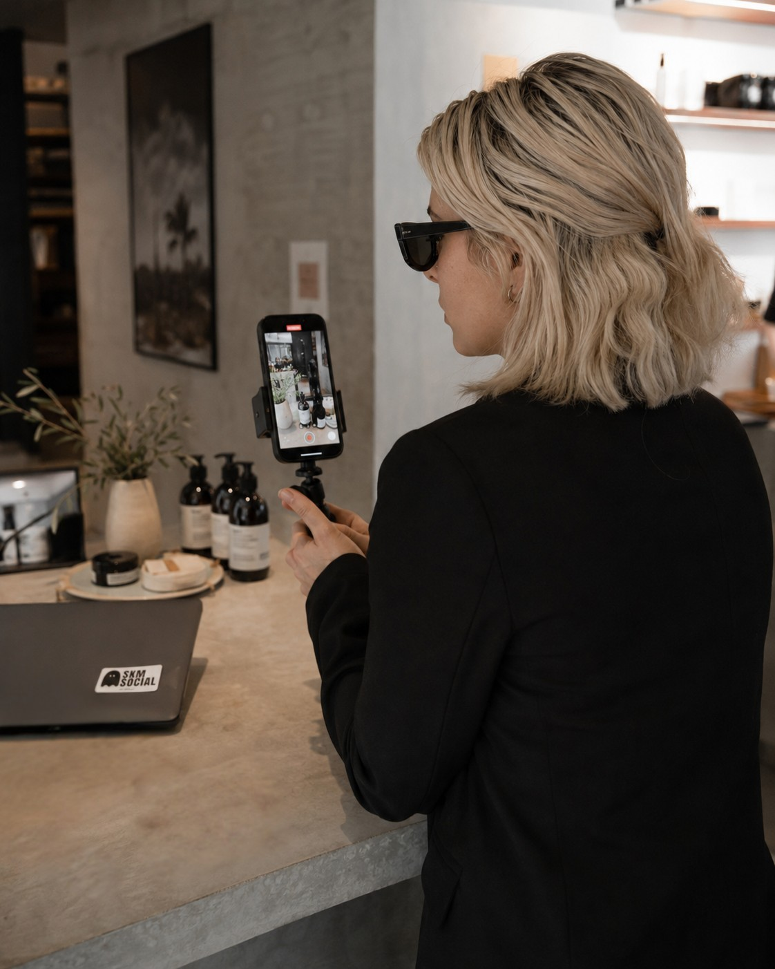 A woman with blonde wavy hair wearing black sunglasses and a black blazer getting content on a selfie stick inside a modern room with a concrete counter, bottles, a plant, and a laptop. Located in Kelowna BC.