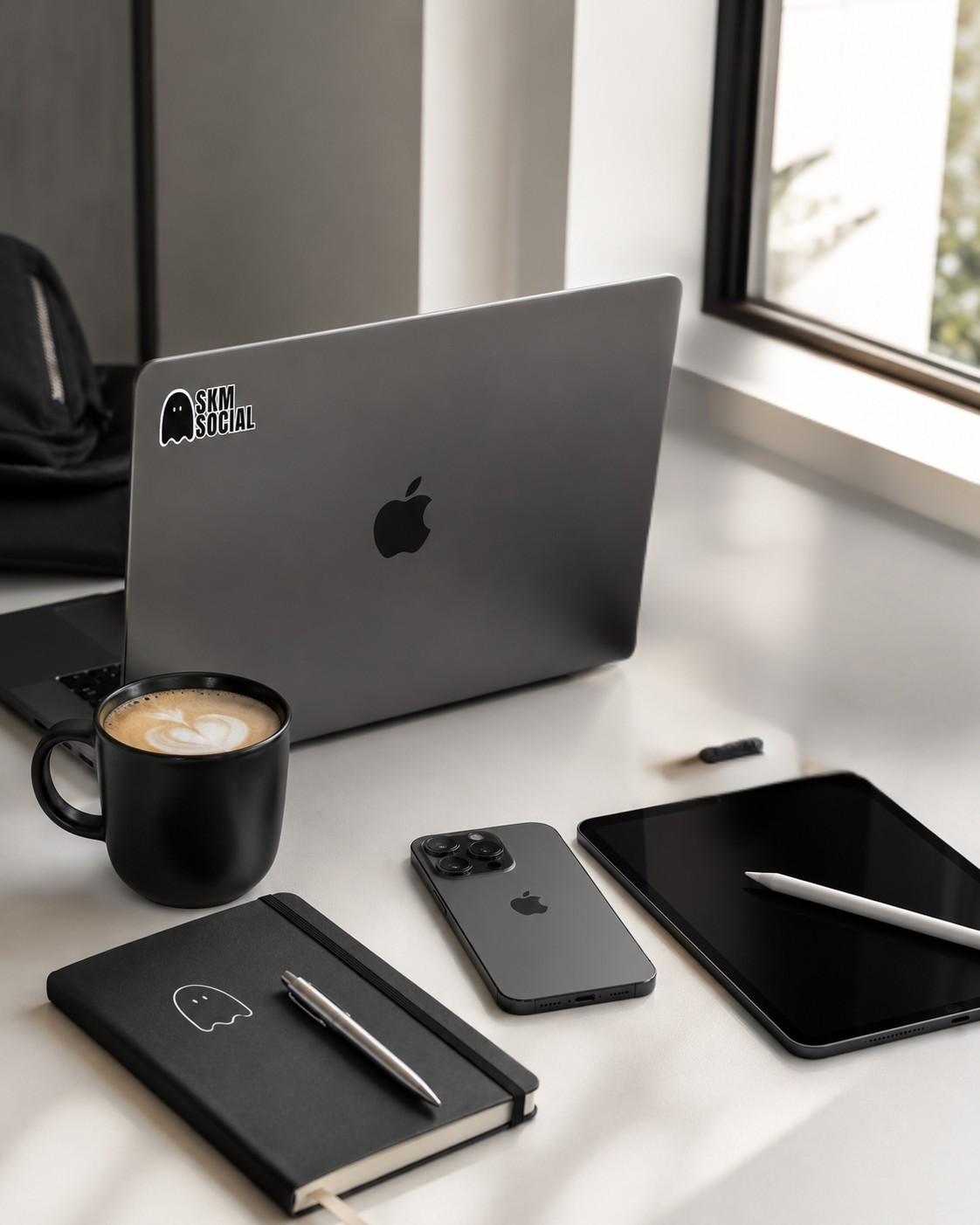 Workspace with a laptop, smartphone, tablet, notebook, pen, coffee mug, and a stylus on a white desk near a window in West Kelowna, British Columbia.