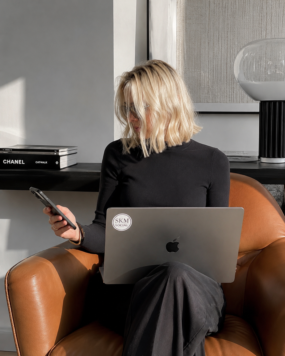 A woman with blonde hair and glasses sitting on a brown leather chair, looking at her phone while a laptop rests on her lap working on a filming brief.