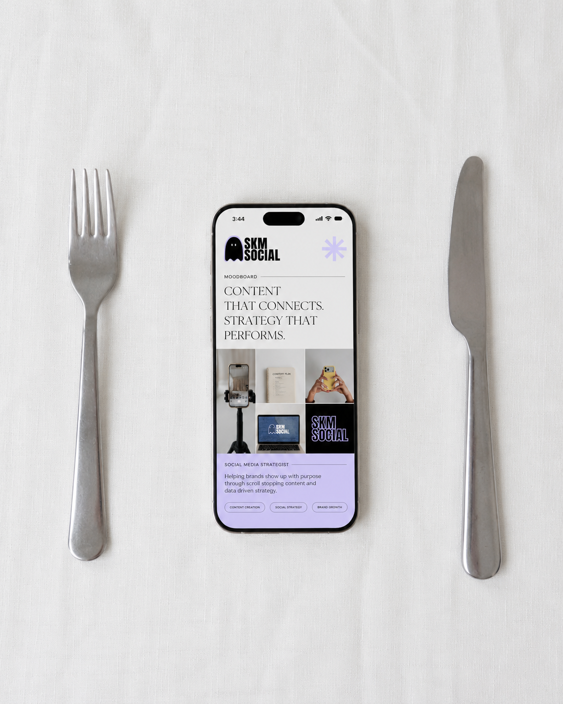 Smartphone on white table between knife and fork, displaying the SKM Social website with text and images about social media content.