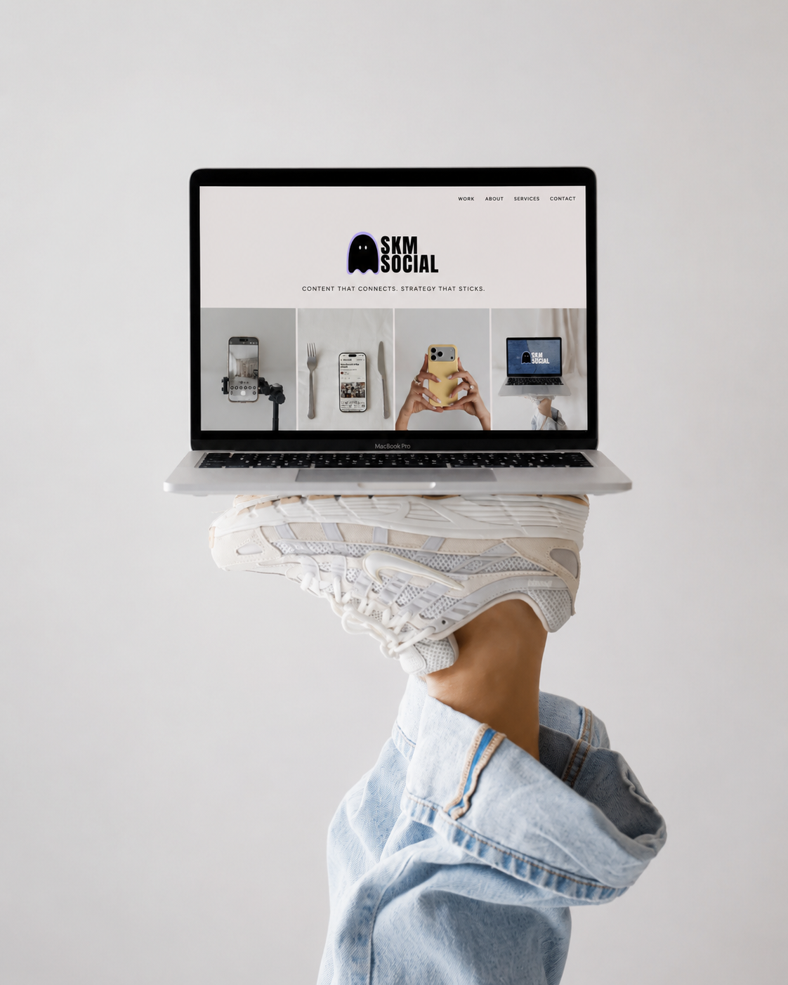 Person wearing white sneakers and light blue jeans holding a MacBook Pro laptop on their foot against a plain white wall promoting filming services for social media.