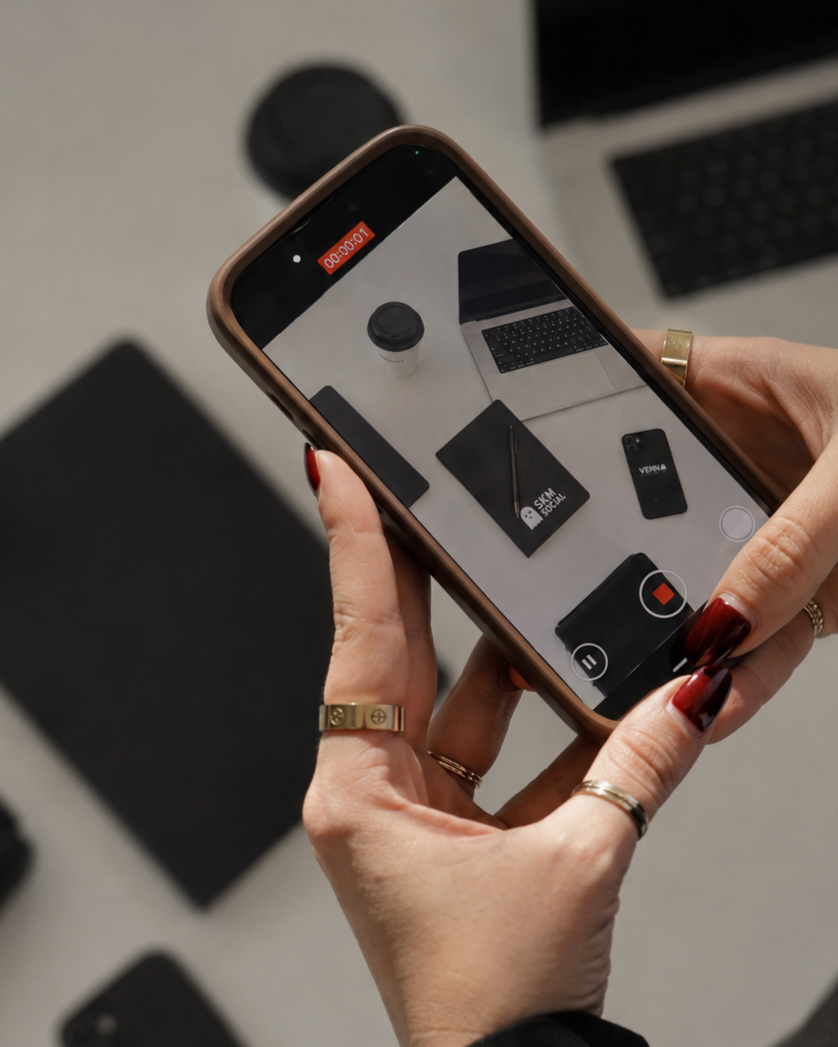 Person taking a photo of a desk setup with a smartphone. The desk holds a coffee cup, a laptop, a drawing tablet, a pen, a smaller black device, and a black wallet.
