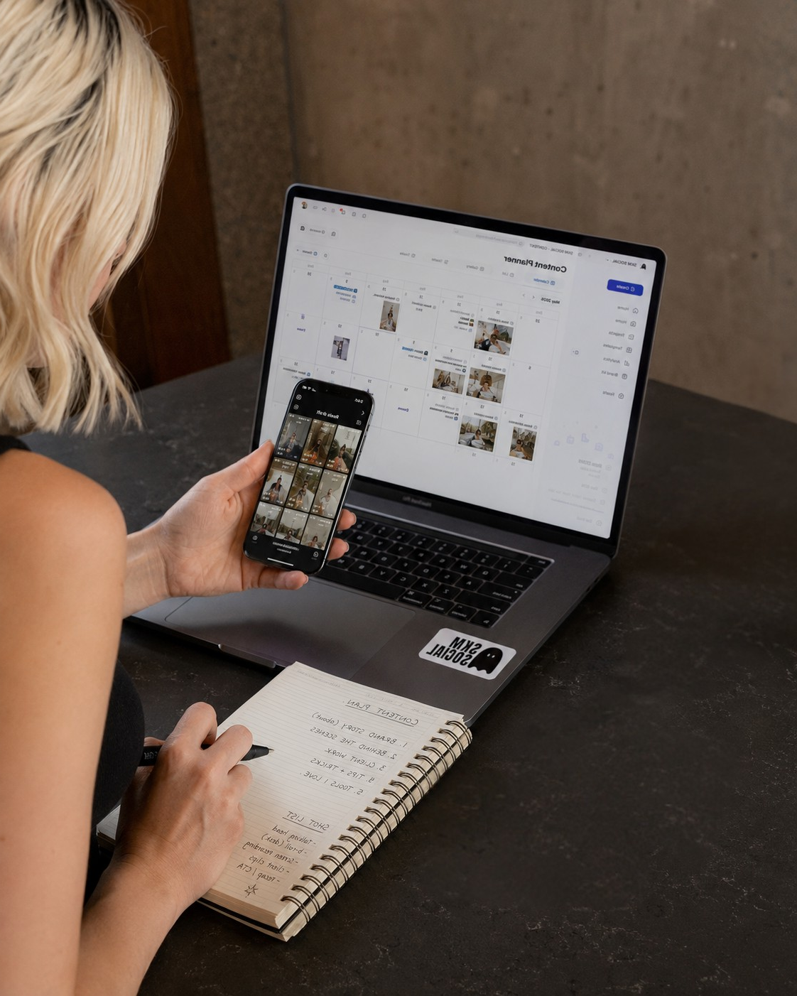 A woman sitting at a dark table with a laptop and a notebook, looking at her phone displaying a grid of photos. The laptop screen shows a calendar with images, and the notebook has handwritten notes about ideas for social media videos.