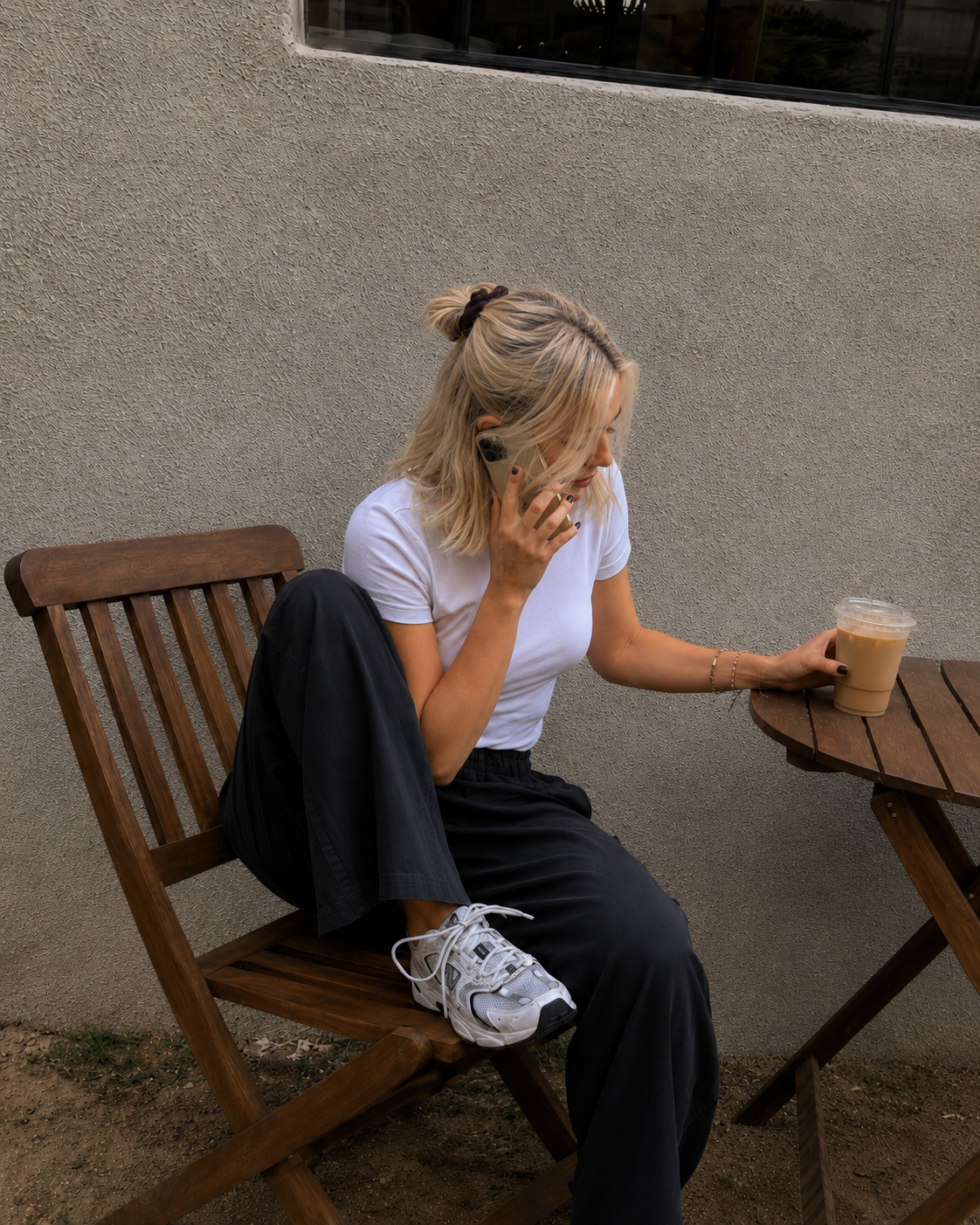 Samm with shoulder-length blonde hair tied with a black hair tie, wearing a white t-shirt and black pants, sitting on a wooden outdoor chair next to a small round table. She is holding a smartphone to her ear and has a large iced coffee on the table.