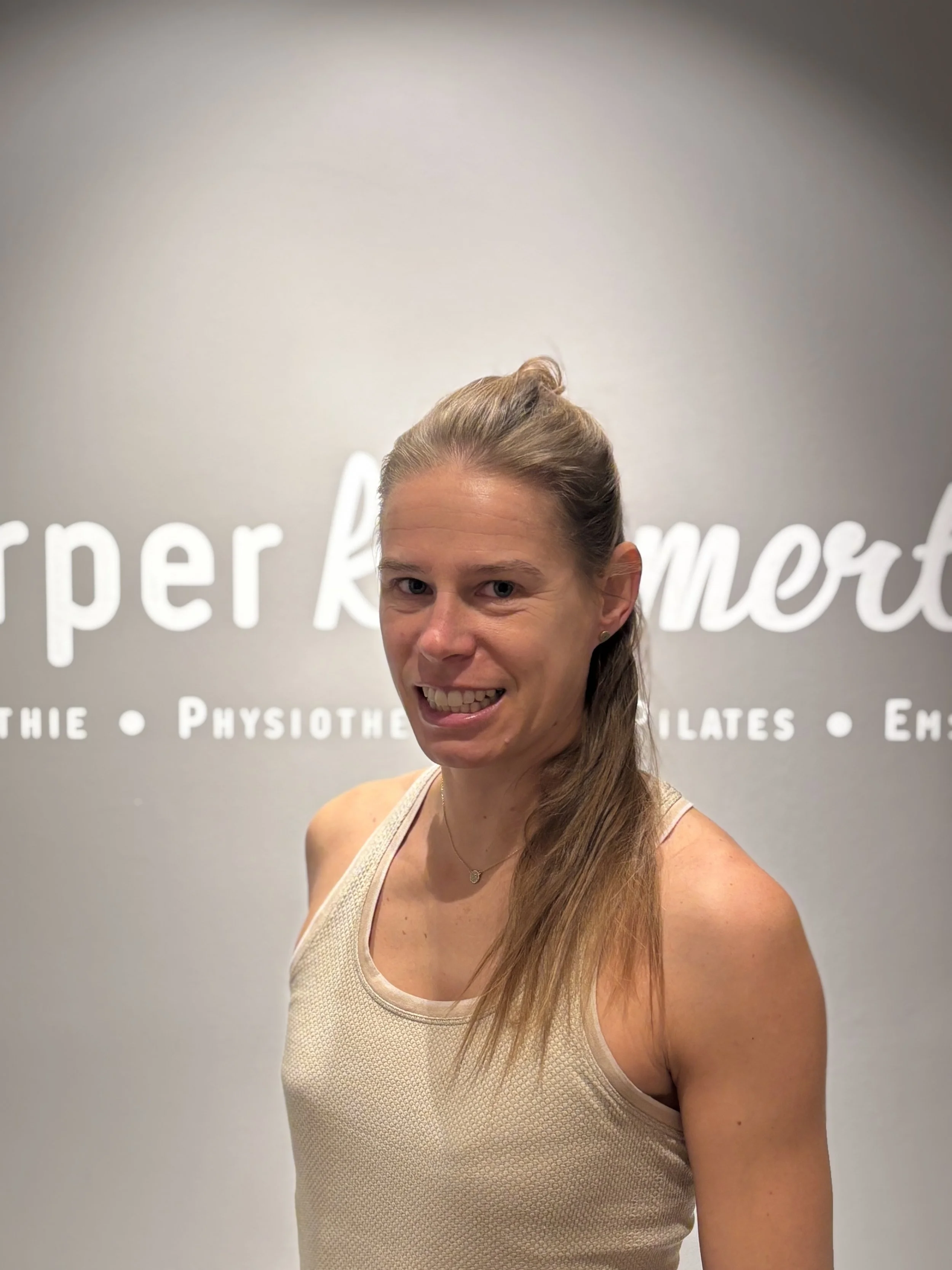 Eine lächelnde Frau steht vor einer grauen Wand mit weißen Schriftzug. Sie trägt ein beiges ärmelloses Top und hat lange braune Haare.