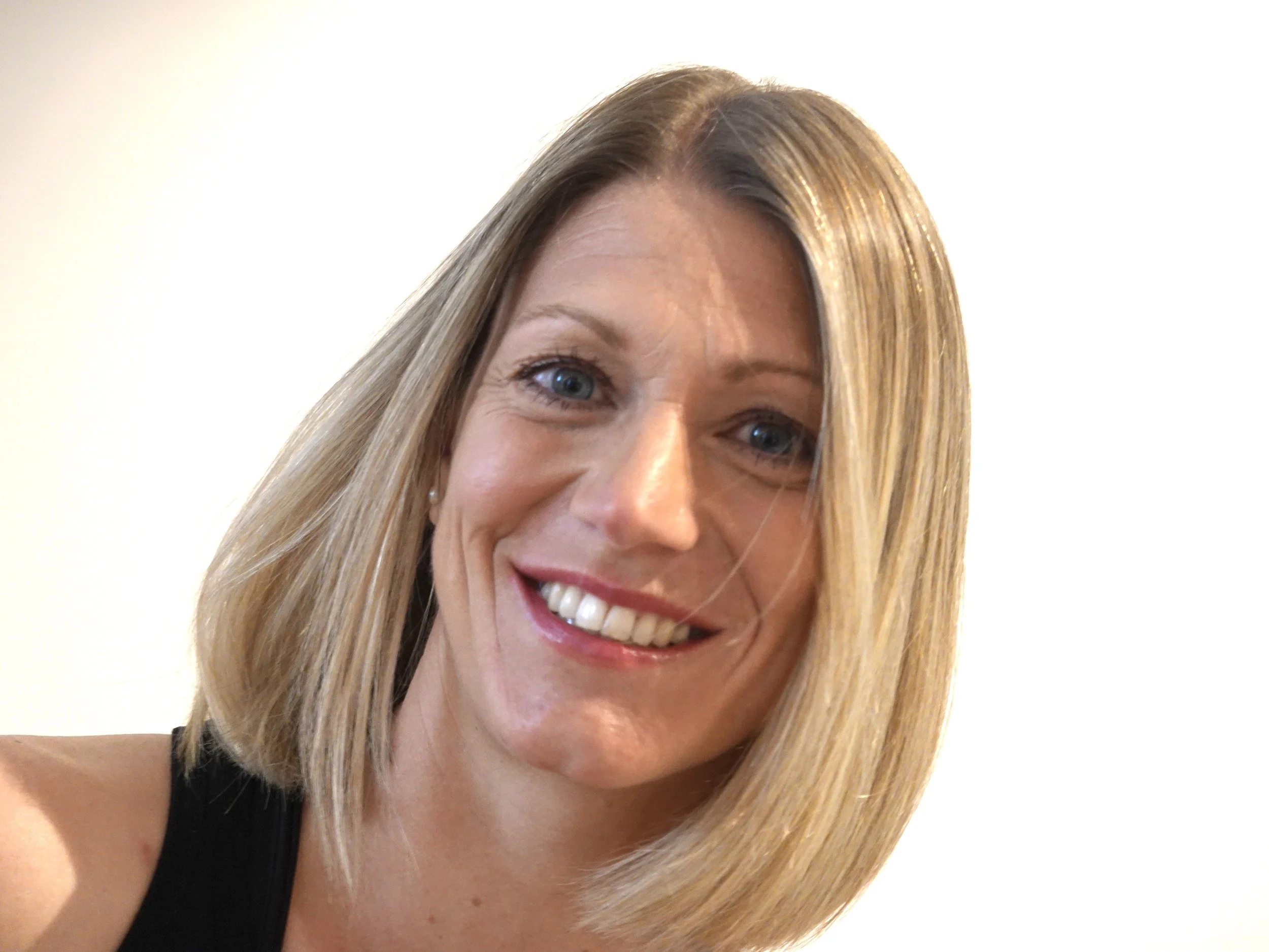 Ein Porträt einer lachenden Frau mit blonden Haaren und blauen Augen, trägt ein schwarzes Oberteil, vor einem hellen Hintergrund.