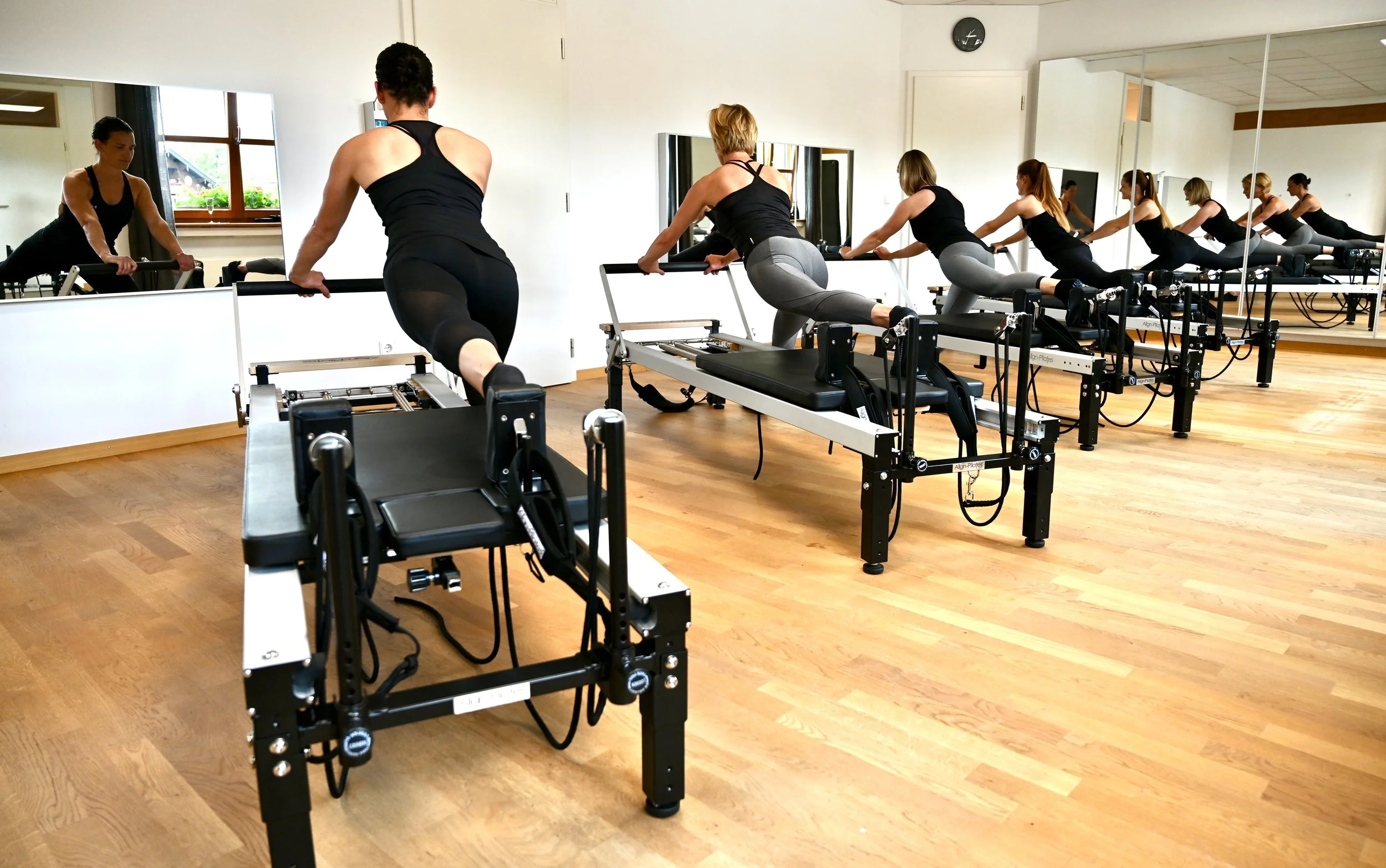 Frauen beim Pilates-Training auf Reformer-Geräten in einem Fitnessstudio mit Holzboden und Spiegeln.
