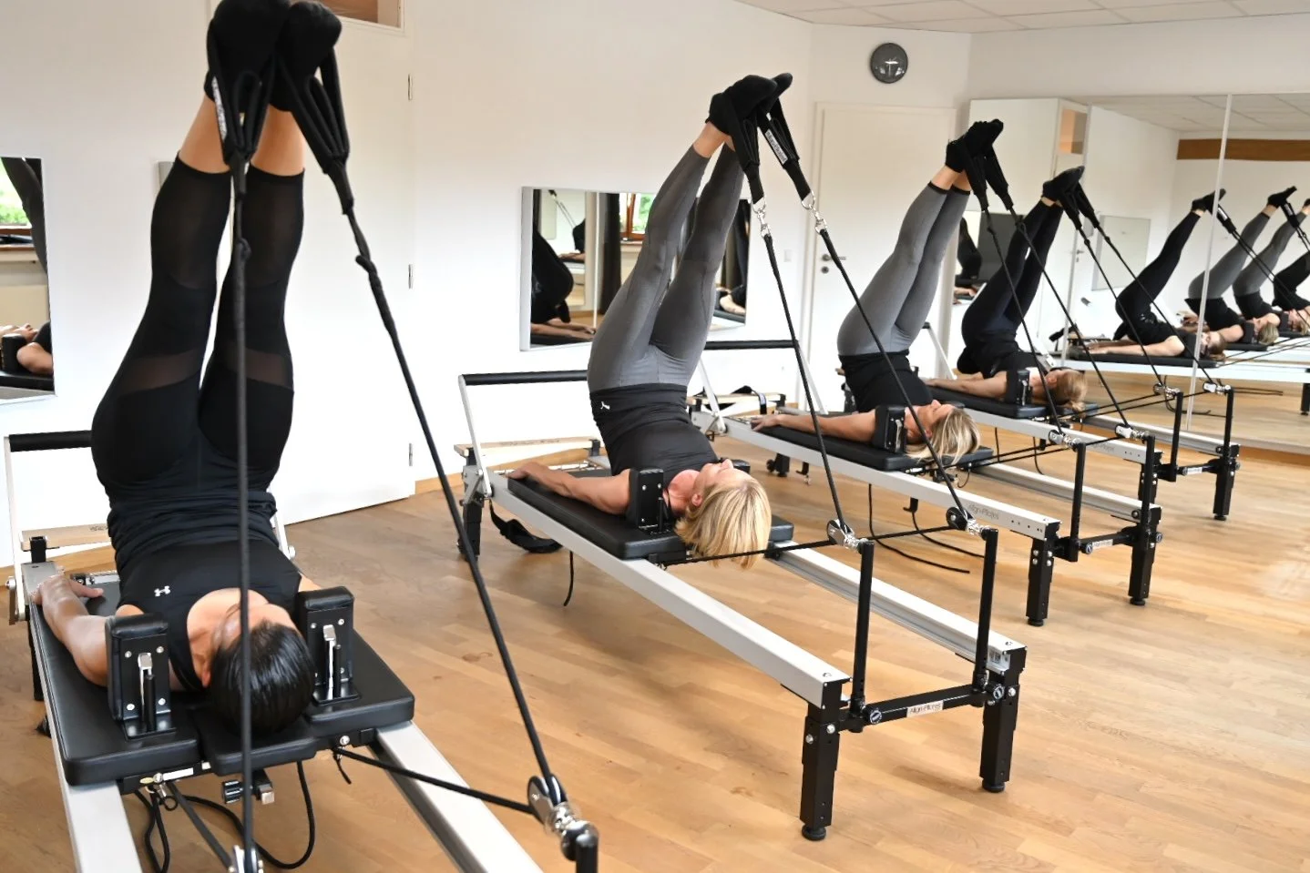 Gruppe von Frauen, die in einem Fitnessstudio auf Pilates-Reformern trainieren, alle liegen auf den Rücken mit gestreckten Beinen und werden durch Trapeze gezogen.