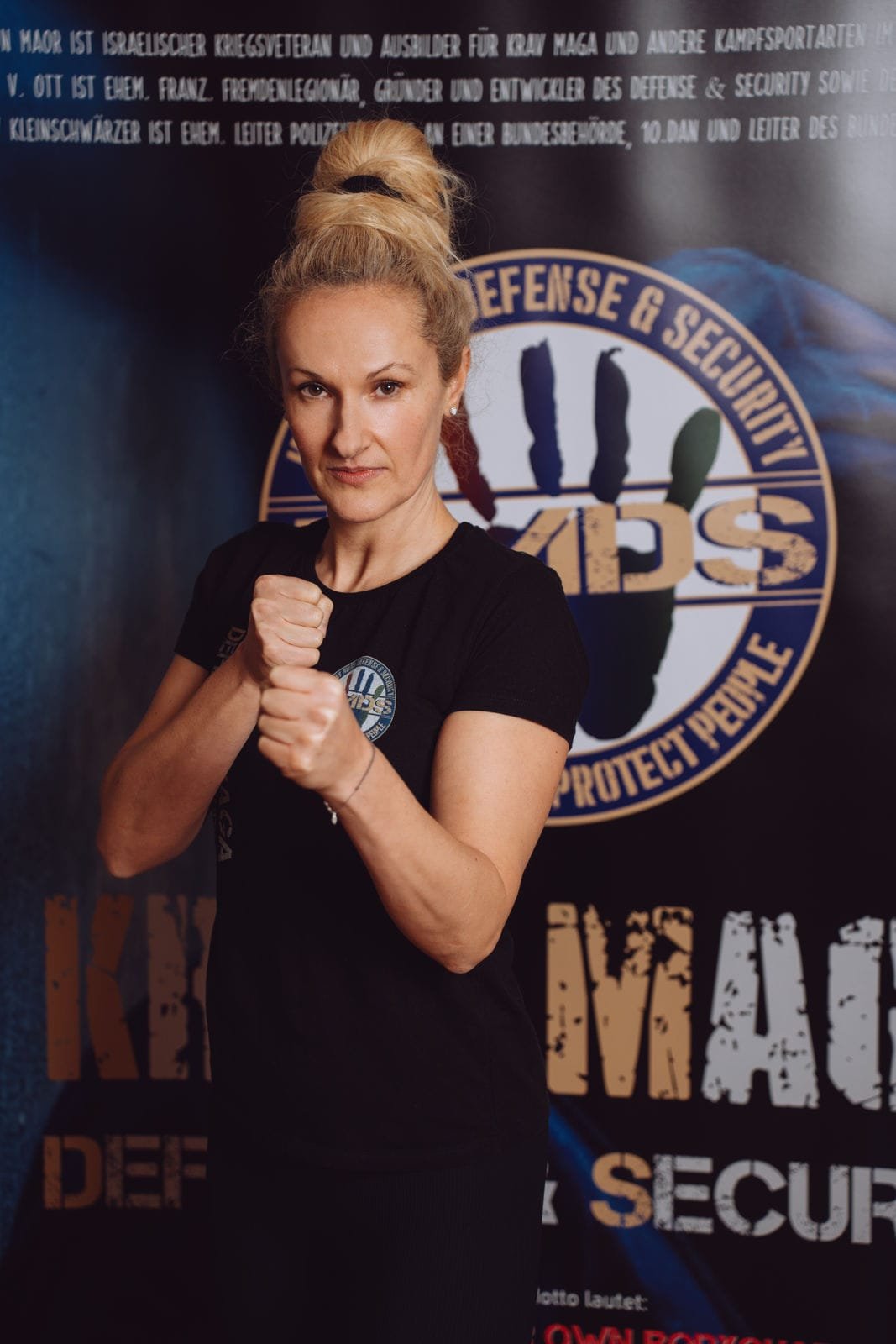 Eine Frau posiert in Kampfsporthaltung vor einem Poster mit dem Text "Defense & Security" und einem Logo, die eine ernsthafte Miene macht.