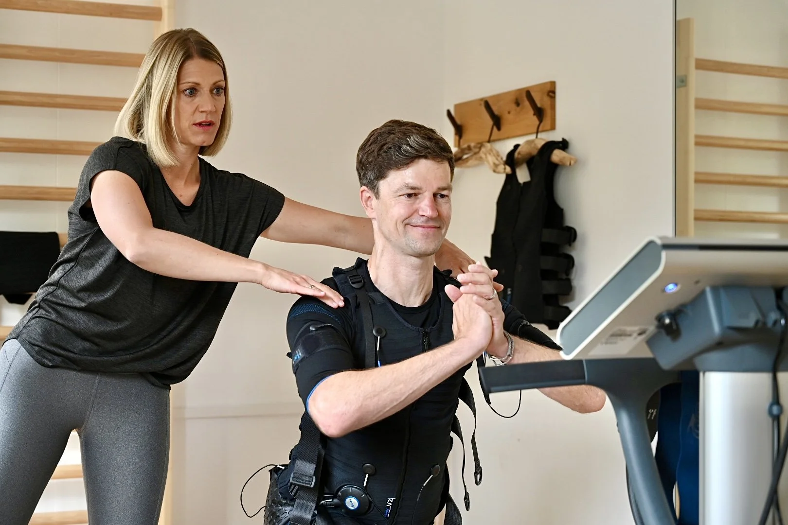 Ein Mann in einem Exoskelett trainiert auf einem Laufband, während eine Trainerin ihm im Fitnessstudio Anweisungen gibt.