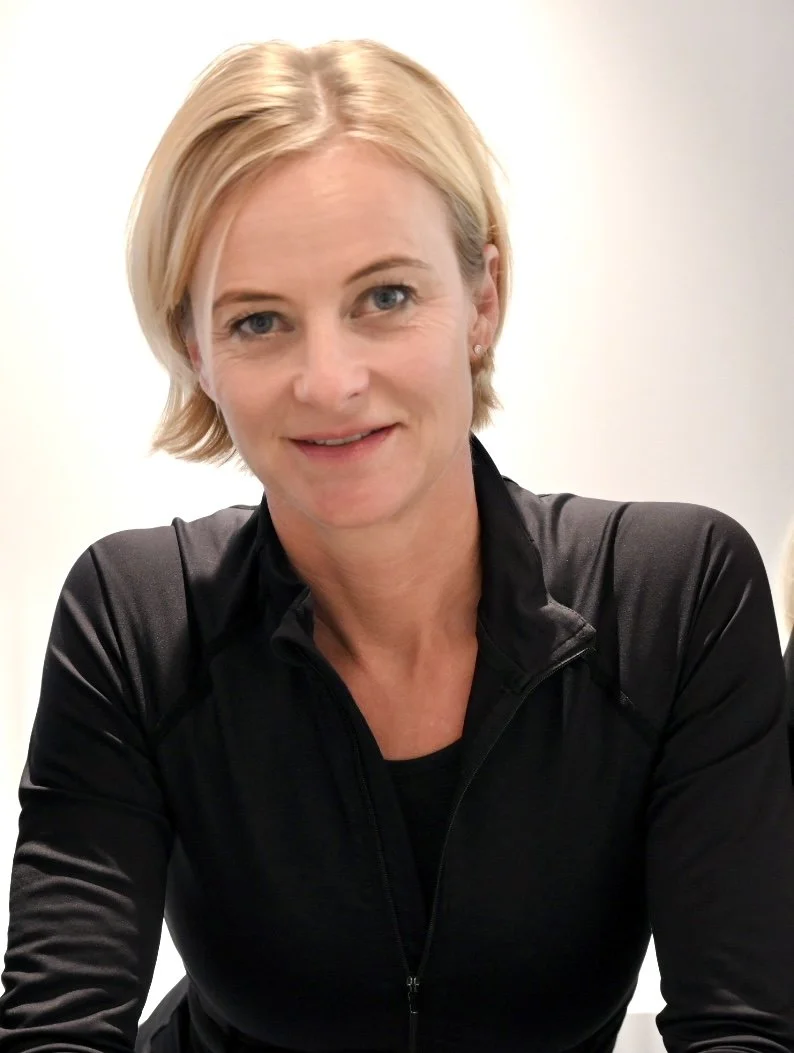 Eine blonde Frau lächelt, trägt eine schwarze Sportjacke und sitzt vor einem weißen Hintergrund.