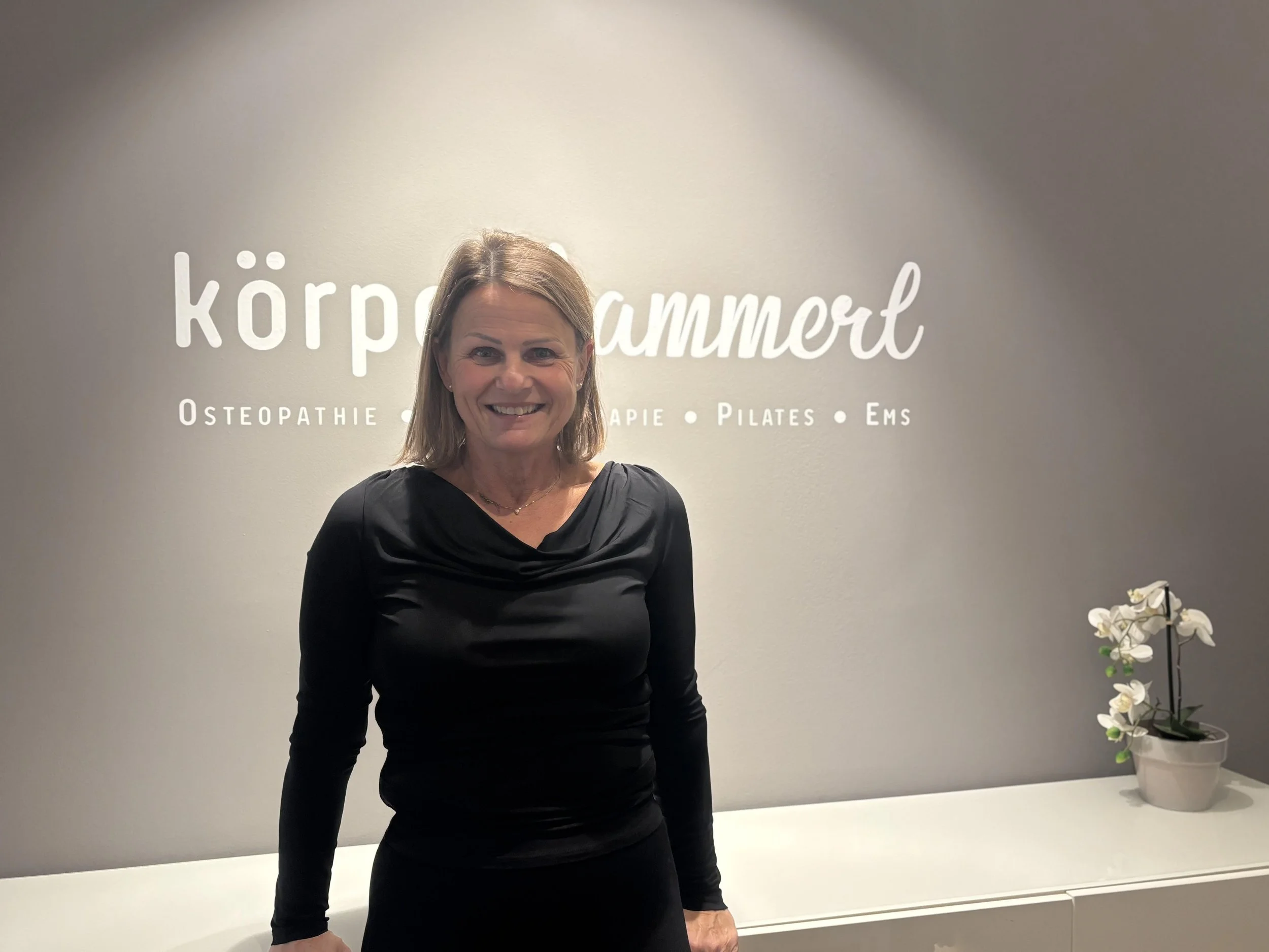Eine lächelnde Frau steht vor einer Wand mit dem Text "körper summaert" und weiteren Worten wie "Osteopathie", "Therapie", "Pilates" und "Ems". Auf der rechten Seite steht eine Topfpflanze.