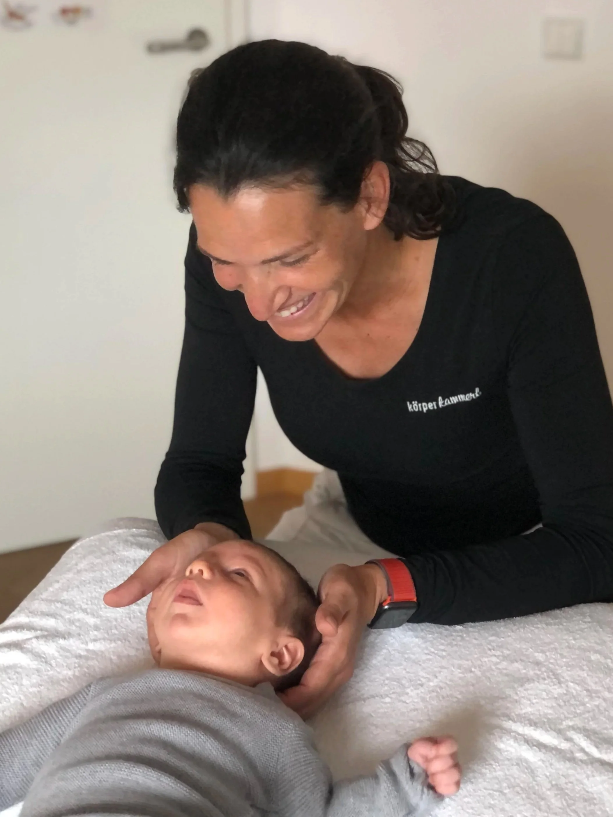 Eine Frau hält einen kleinen Jungen auf einem Behandlungstisch, beide lachen und schauen sich an.