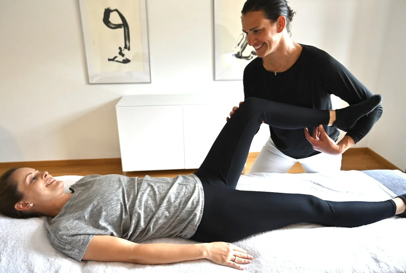 Eine Therapeutin führt eine Physiotherapie an einer Patientin durch, die auf einer Behandlungsliege liegt und ihre Beine hebt.