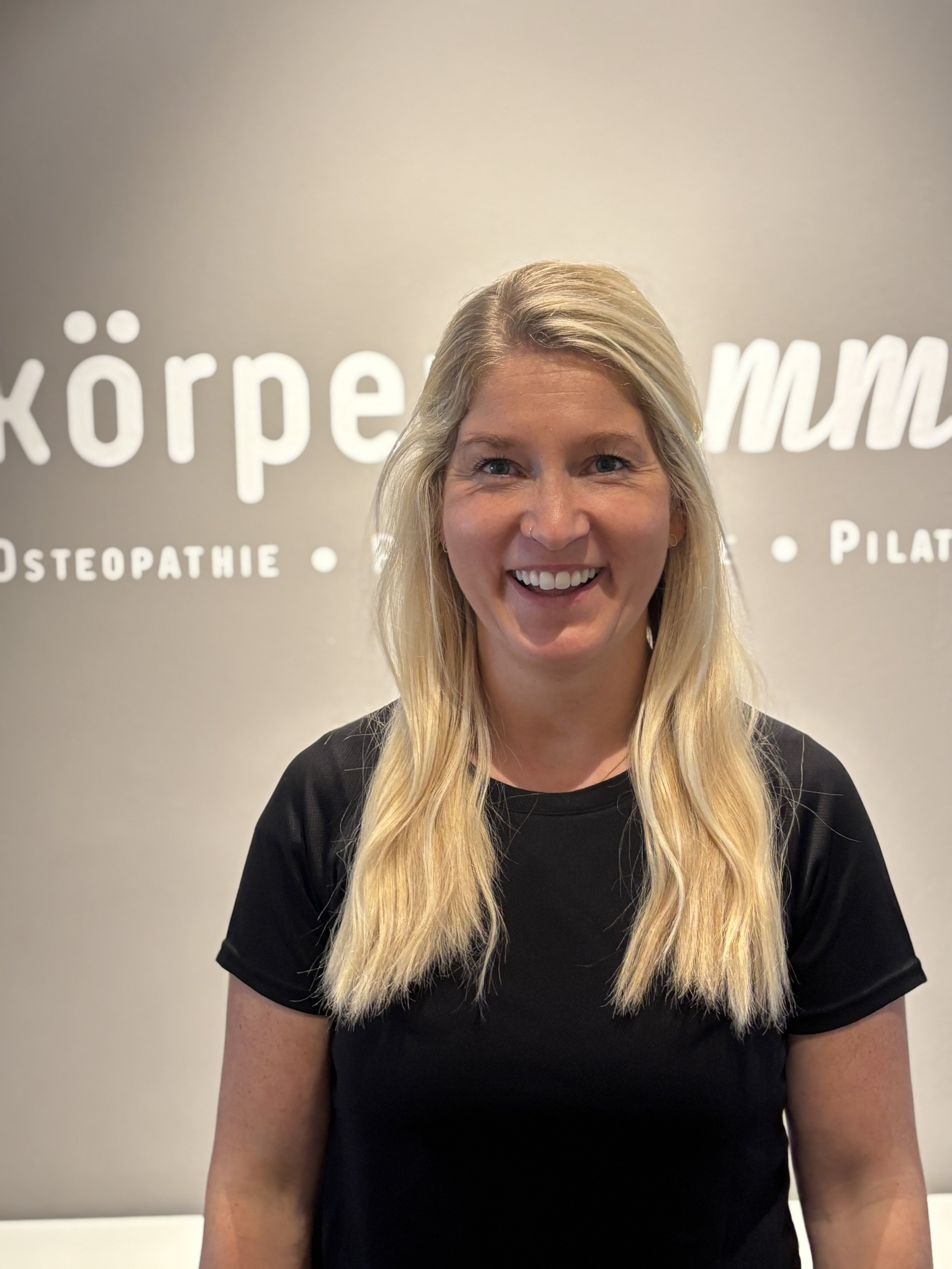 Ein lächelnder blonde Frau in schwarzener T-Shirt vor einer grauen Wand mit weißem Text.