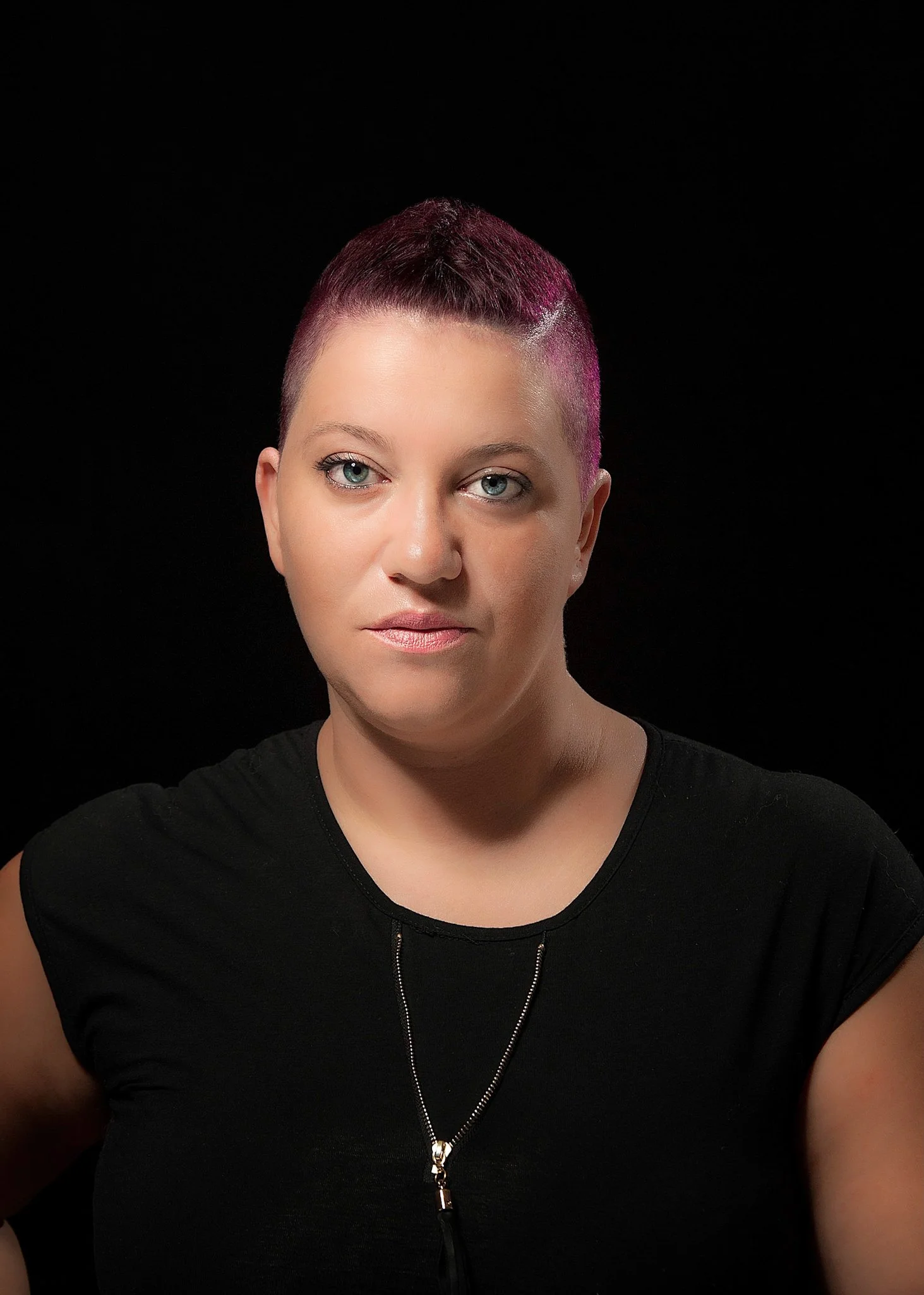 Portrait of a woman with short, dark pink hair, light skin, green eyes, wearing a black top and a necklace, against a black background.