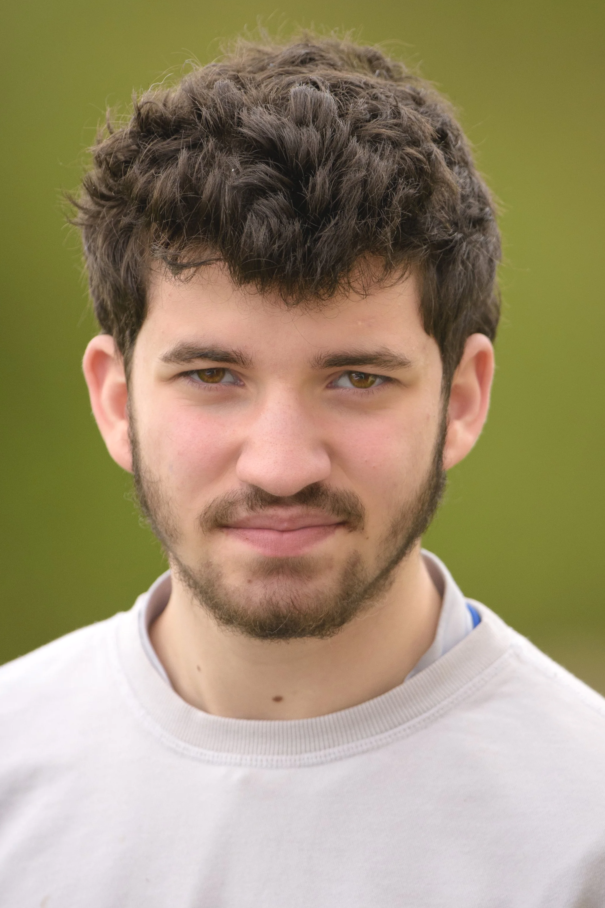 Close-up of a young man with short, dark, curly hair and a beard, wearing a light-colored shirt, outdoors with a blurred green background.