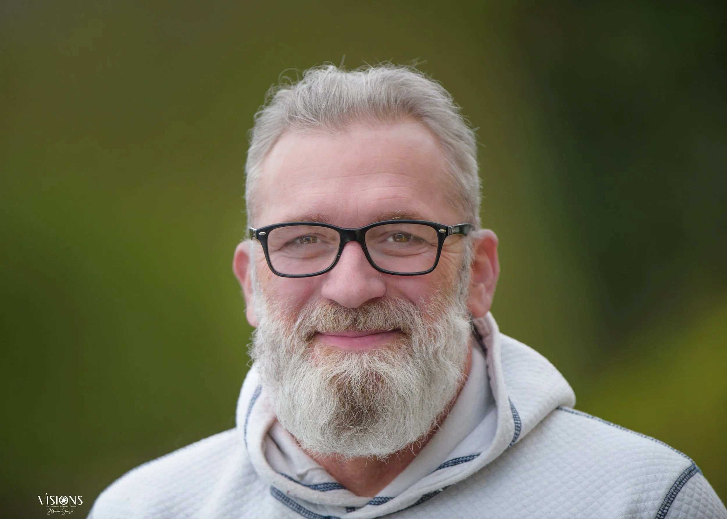 A smiling man with gray hair and beard, wearing glasses and a light-colored hoodie outdoors.
