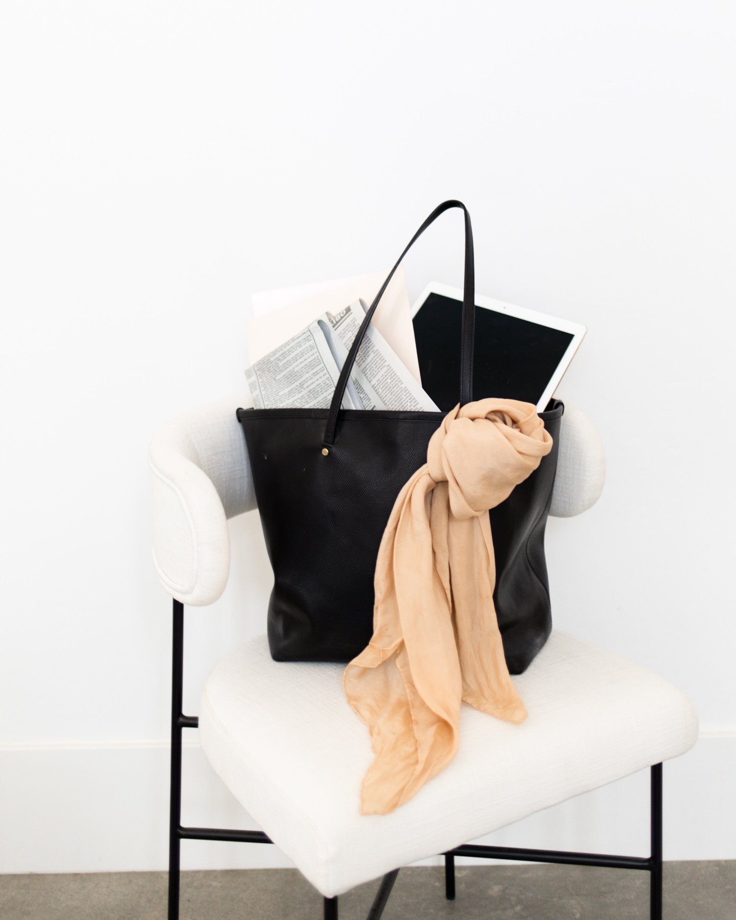 A white armchair with a black arch and a black bag hanging on it. The bag contains papers, an electronic tablet, and a beige scarf.