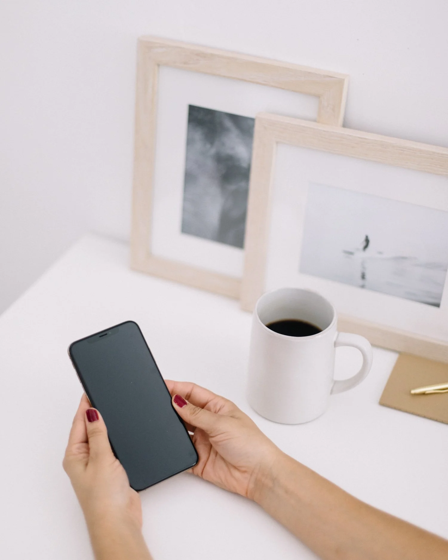 A person with red nail polish holding a dark smartphone over a white table with two framed pictures, a white mug filled with coffee, a notebook, and a pen.