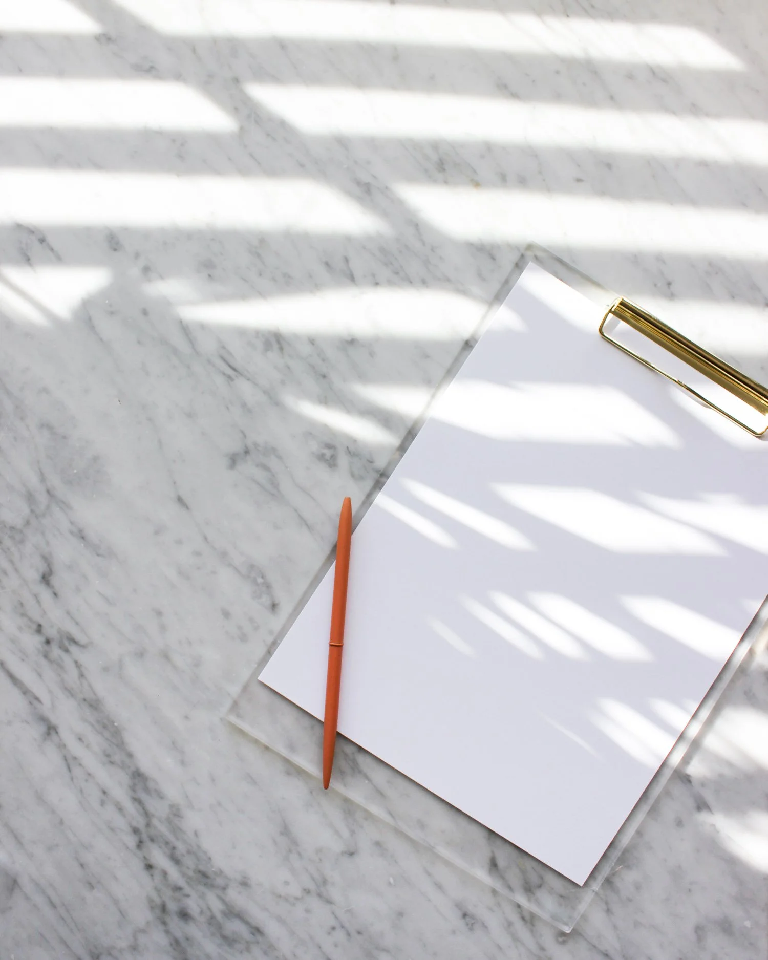 A white sheet of paper with an orange pencil and a gold pen on top, placed on a marble surface with sunlight creating shadows.